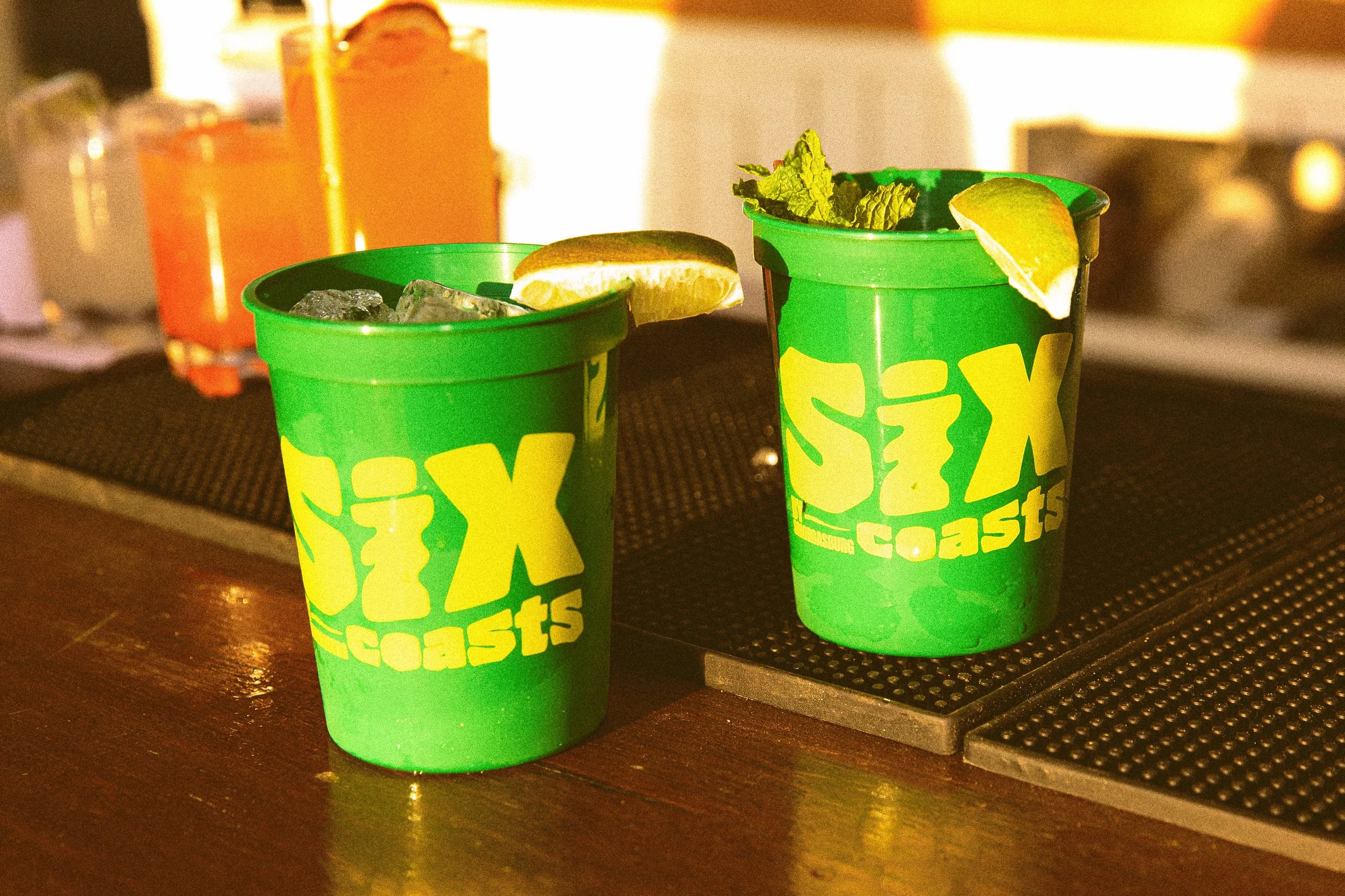 wo signature green Six Coasts cocktails with ice, lime wedges, and a sprig of mint, served on a black bar mat. The background features other orange drinks.