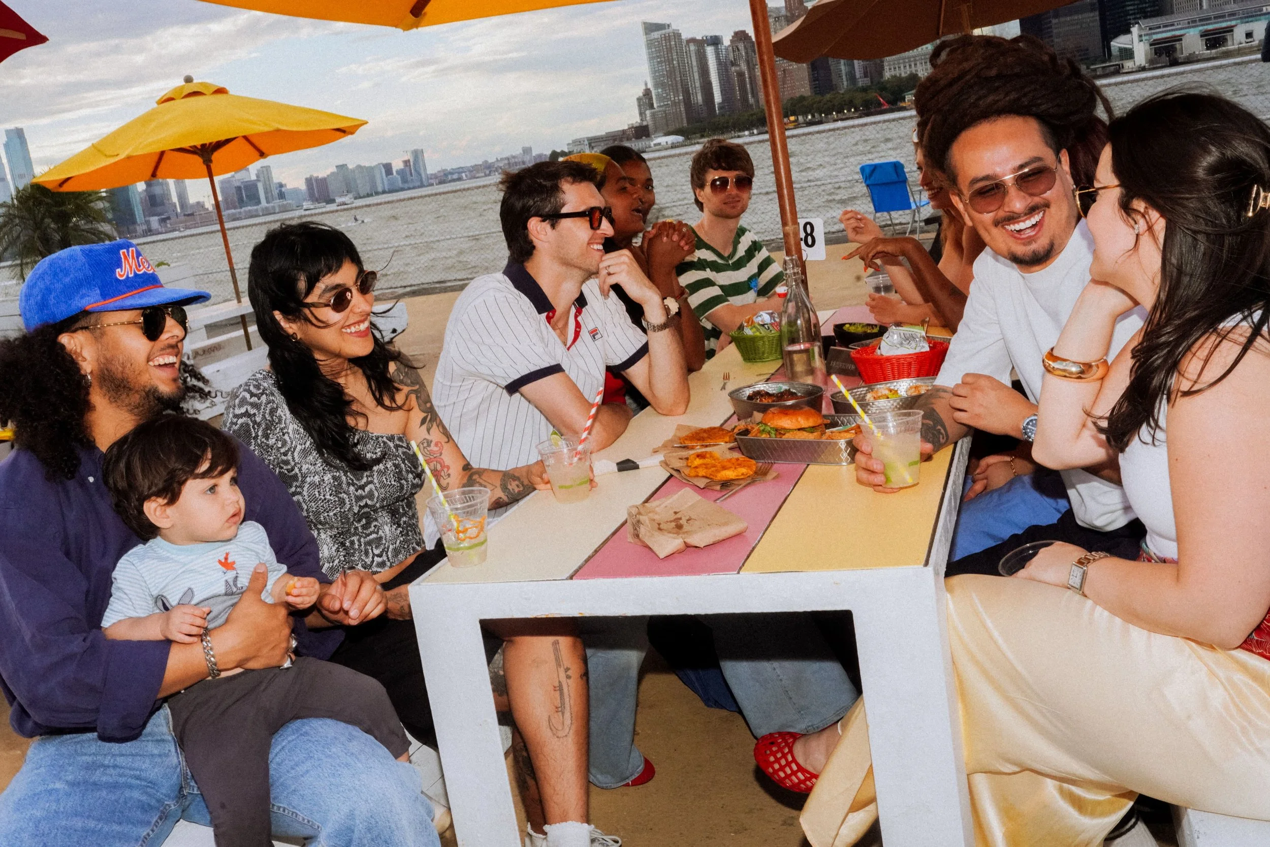 Group of friends enjoying drinks and food at Six Coasts outdoor picnic tables on Governors Island with views of the waterfront and Manhattan skyline.