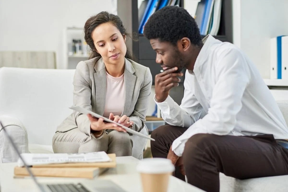 Two professionals, a woman and a man, sitting on a sofa and discussing work documents in an office, with a laptop and coffee cup on the table in front of them.