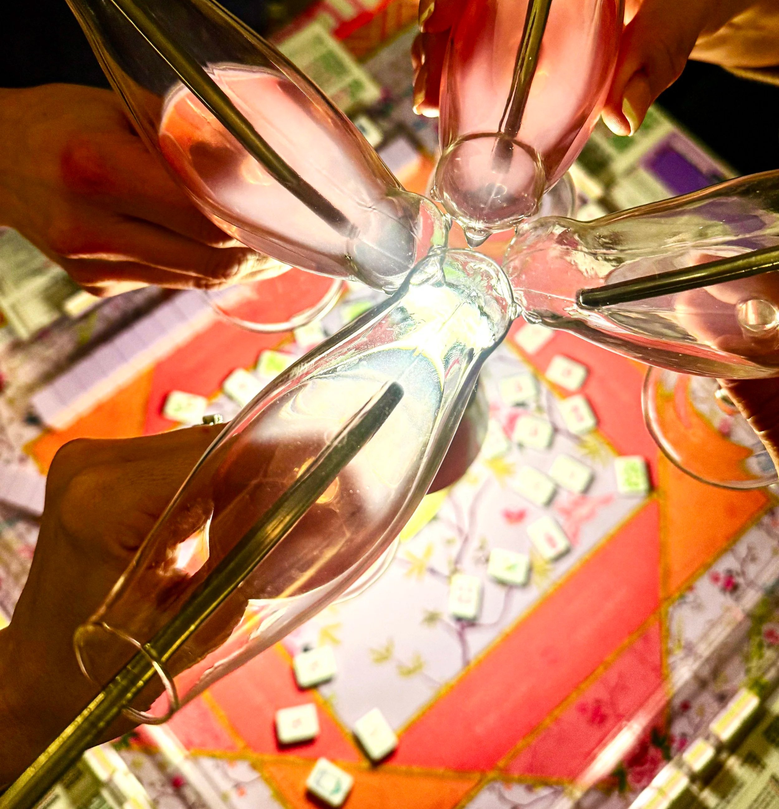 A top-down view of five people clinking empty glass champagne flutes together in a toast. The background shows a table with various objects, possibly a game or decorations, under warm lighting.