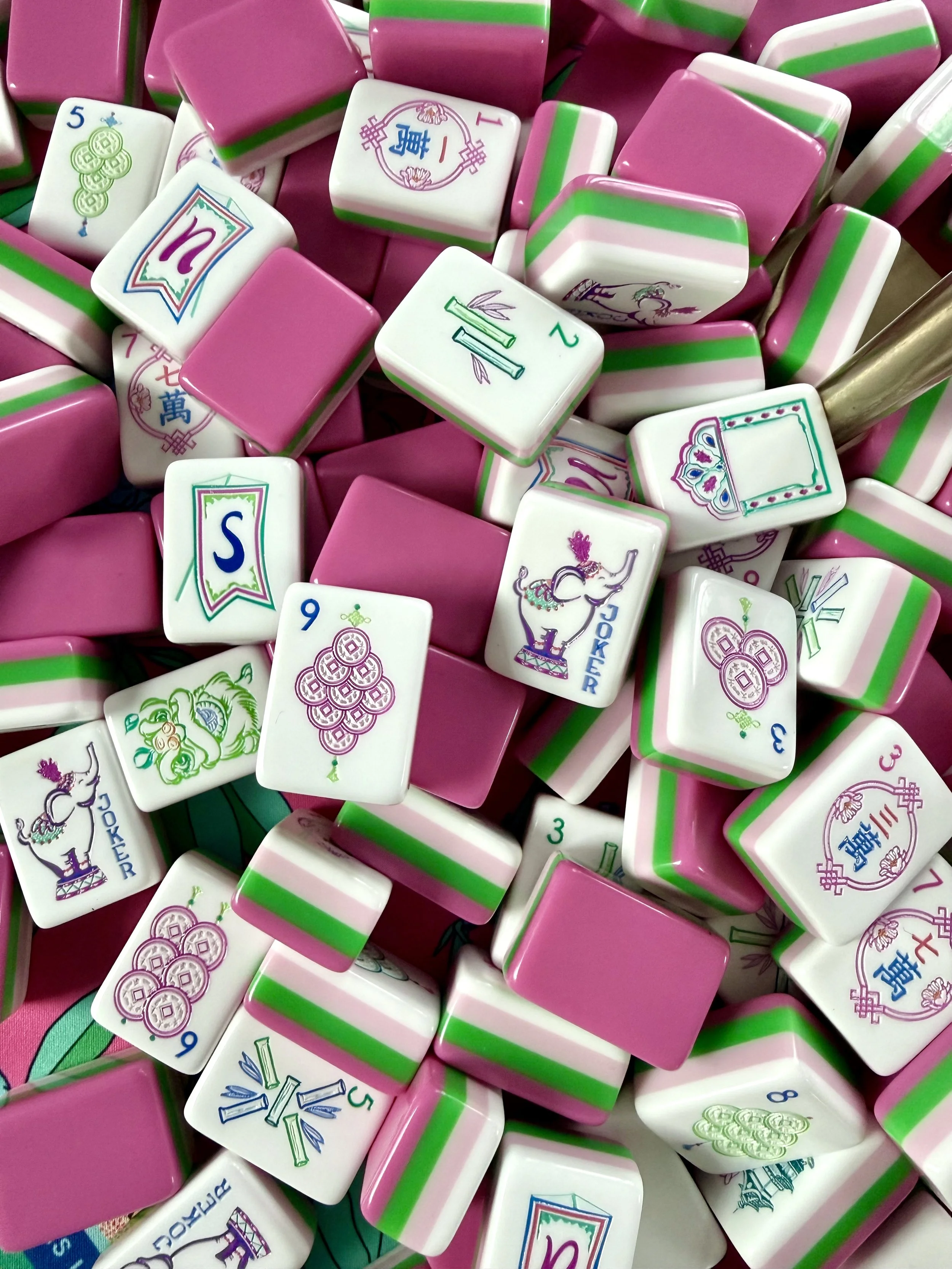 A collection of Mahjong tiles, predominantly white with some pink, featuring various symbols, characters, and illustrations, including elephants and stacks of coins, with some tiles having pink stripes.