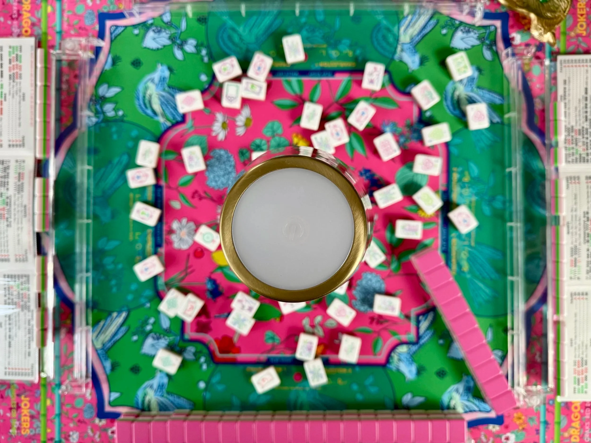 Top-down view of a colorful board game with a central round container of white candles, surrounded by small rectangular tiles and a pink plastic collector tray.