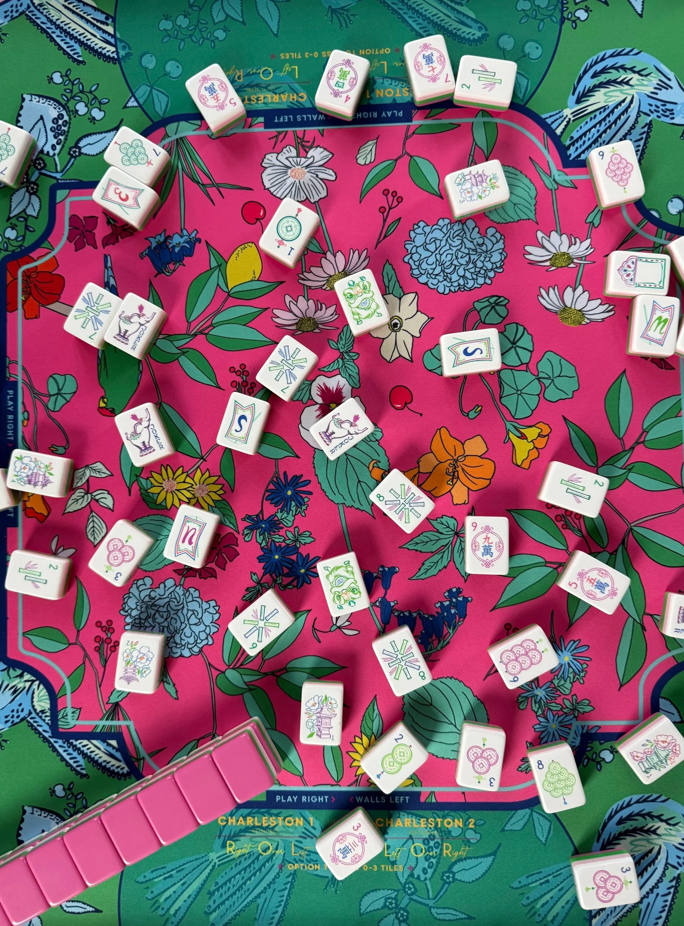 Close-up of a Mahjong game setup with white tiles featuring colorful Chinese characters and symbols, on a vibrant, floral-patterned game mat.