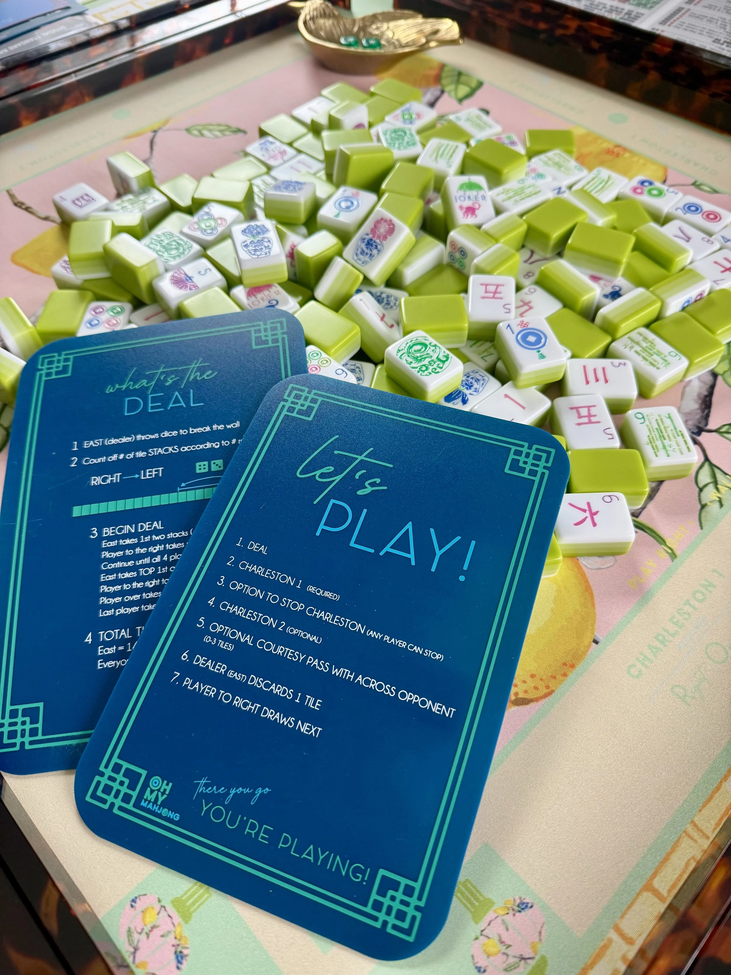 Mahjong game setup with a pile of green and white Mahjong tiles and two blue instruction cards on a colorful Mahjong-themed playmat.