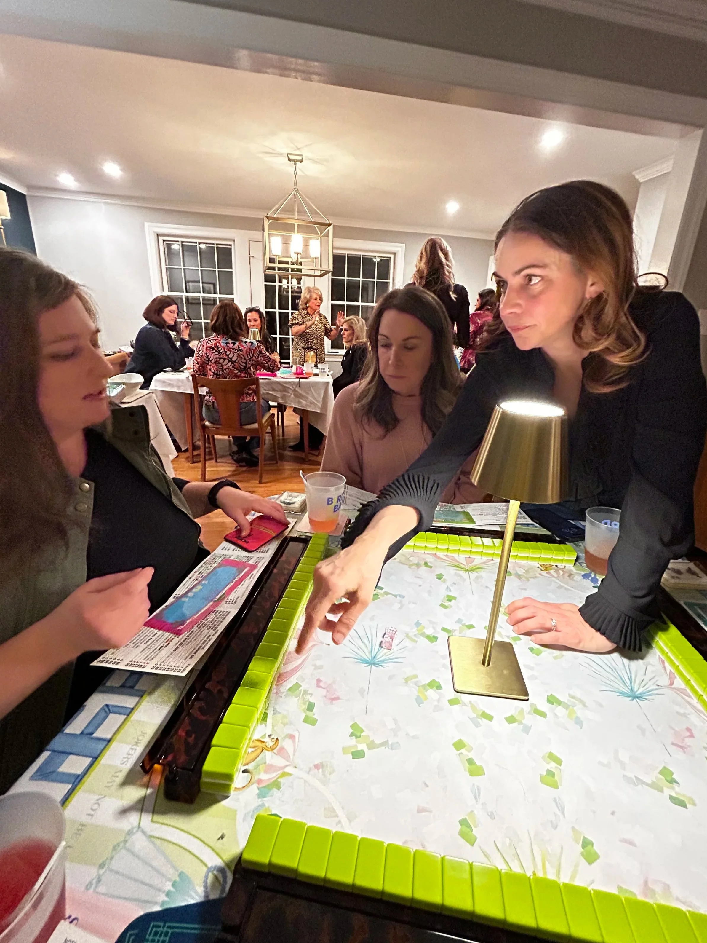 A group of women playing a game of Mahjong around a table with green tiles, a lamp, and various cards, in a well-lit dining room with other women sitting at a larger table in the background.
