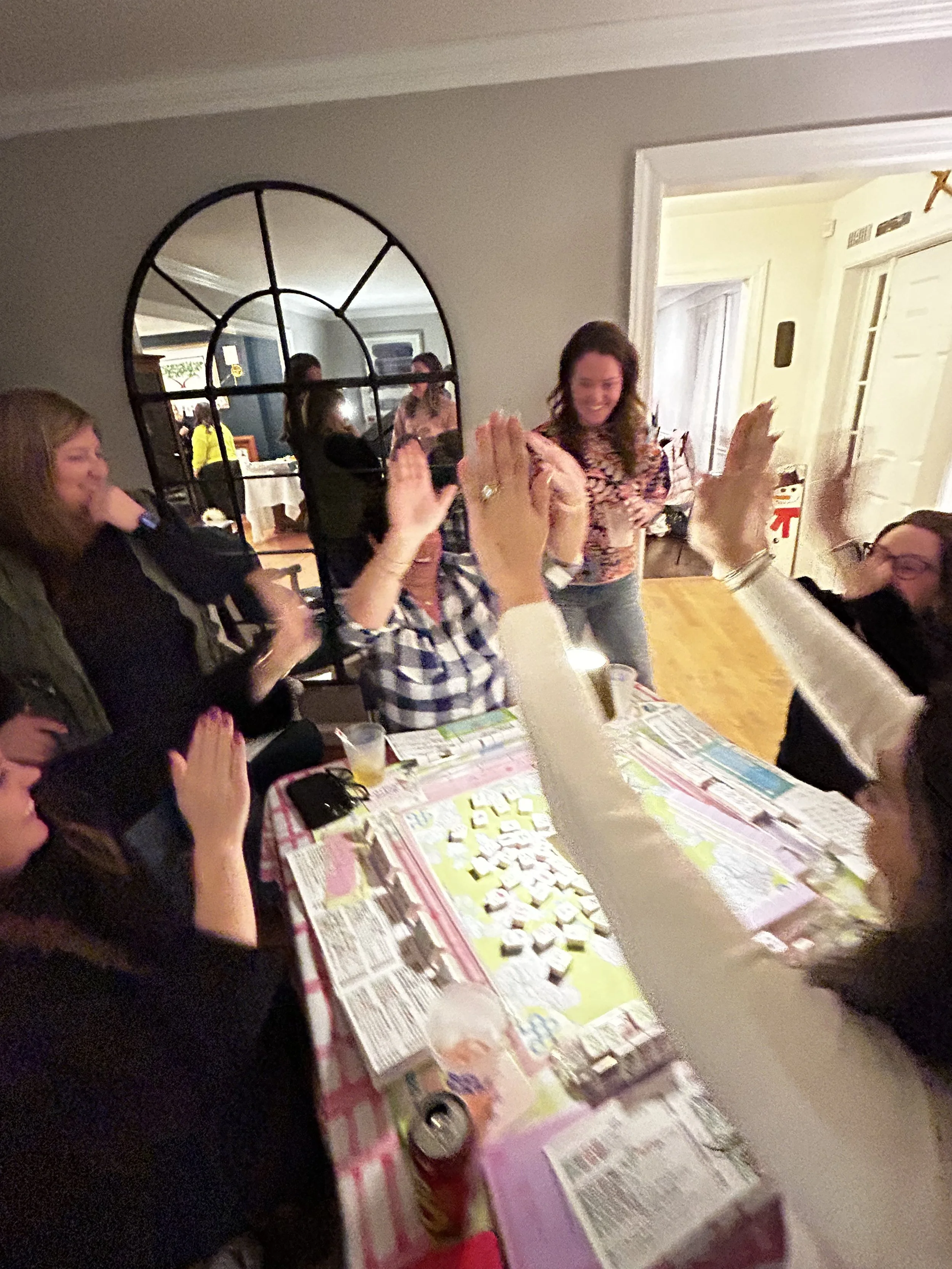 Group of people playing a game at a table, some raising their hands for a high five, in a cozy indoor setting with a mirror and doorway visible in the background.
