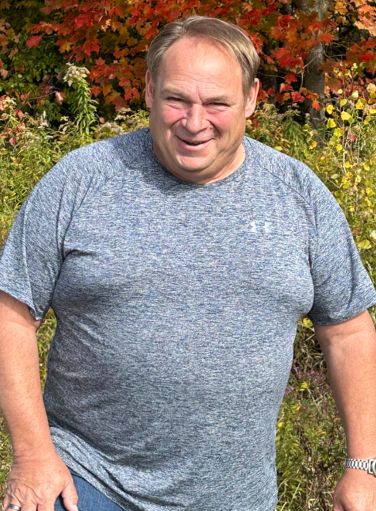 A smiling man in a gray heather T-shirt stands outdoors in front of autumn-colored foliage.