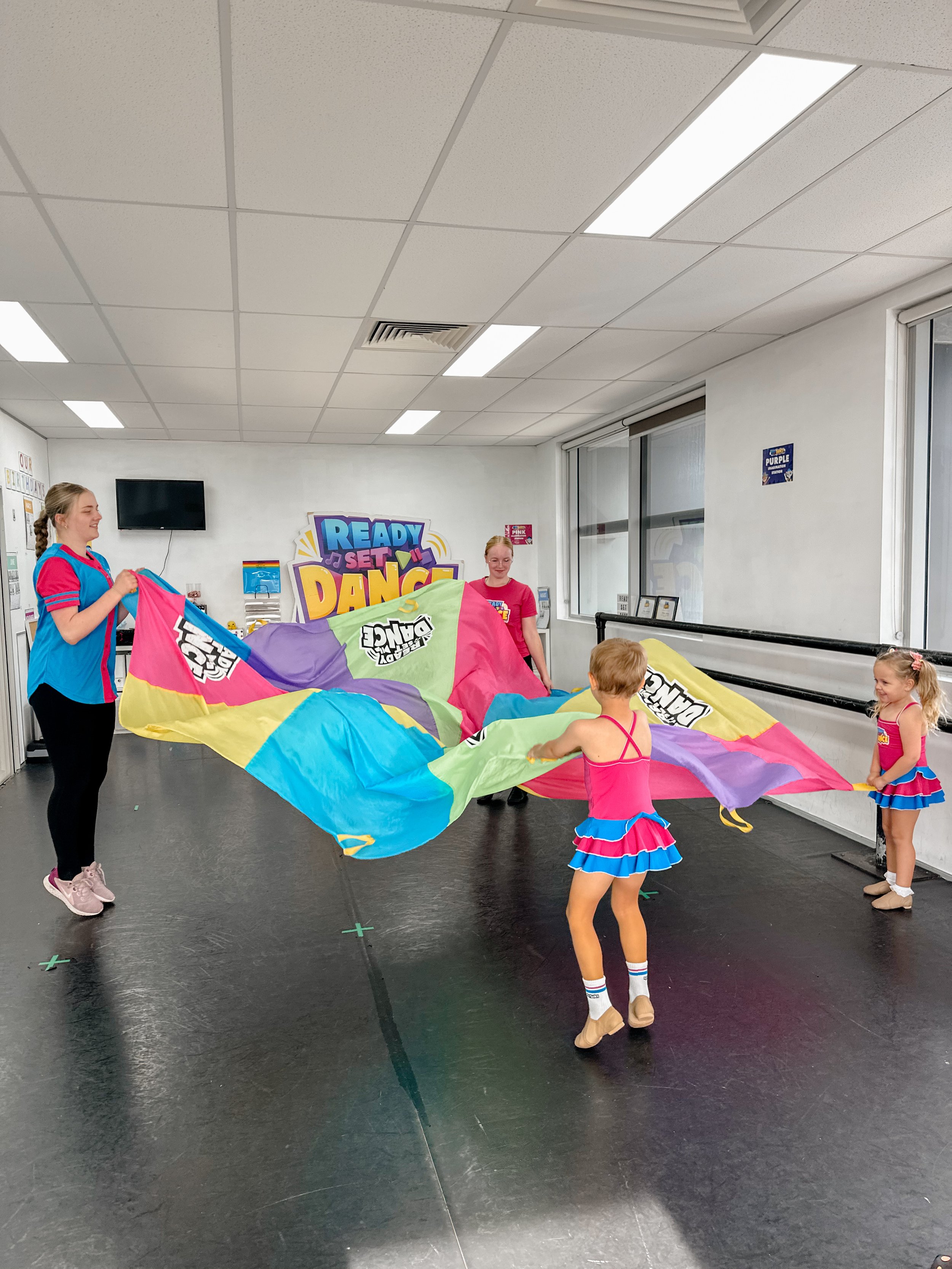 Children and two adults playing with a colorful parachute in a dance studio with a sign that says "Ready Set Dance" in the background.
