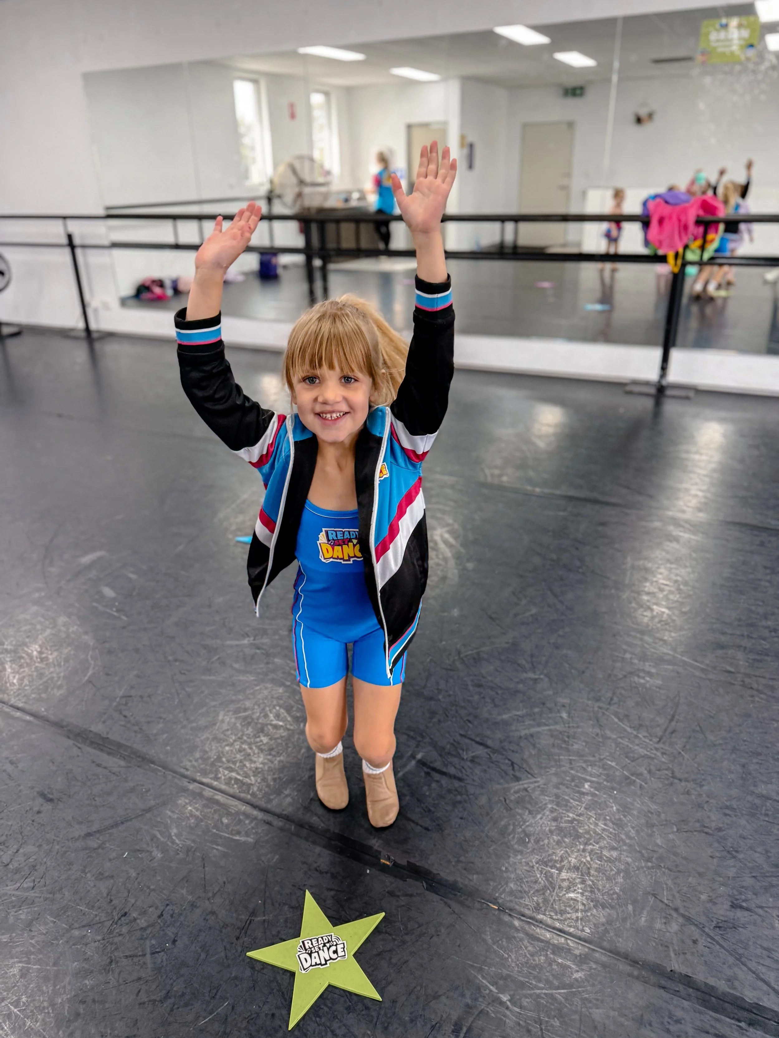 A young girl with blonde hair in dance attire, standing on a dance studio floor with her arms raised in the air, smiling, with a star-shaped award on the floor that says 'Ready to Dance'.
