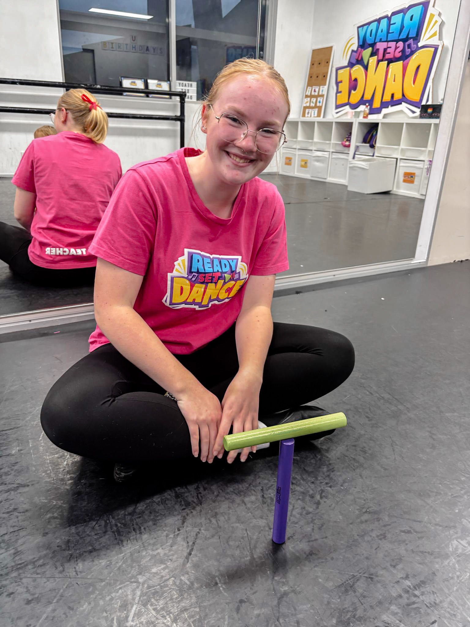 A smiling young woman with red hair, glasses, and a pink T-shirt sitting cross-legged on the floor in a dance studio, balancing a green and purple foam stick on her lap.
