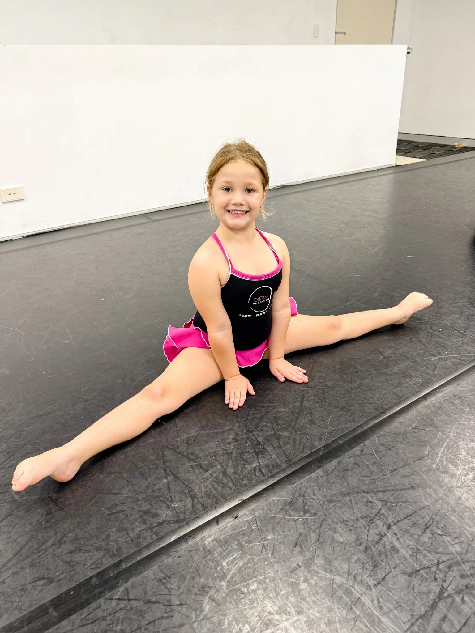 A young girl in a black and pink cheerleading outfit sitting on the floor in a split position, smiling at the camera.