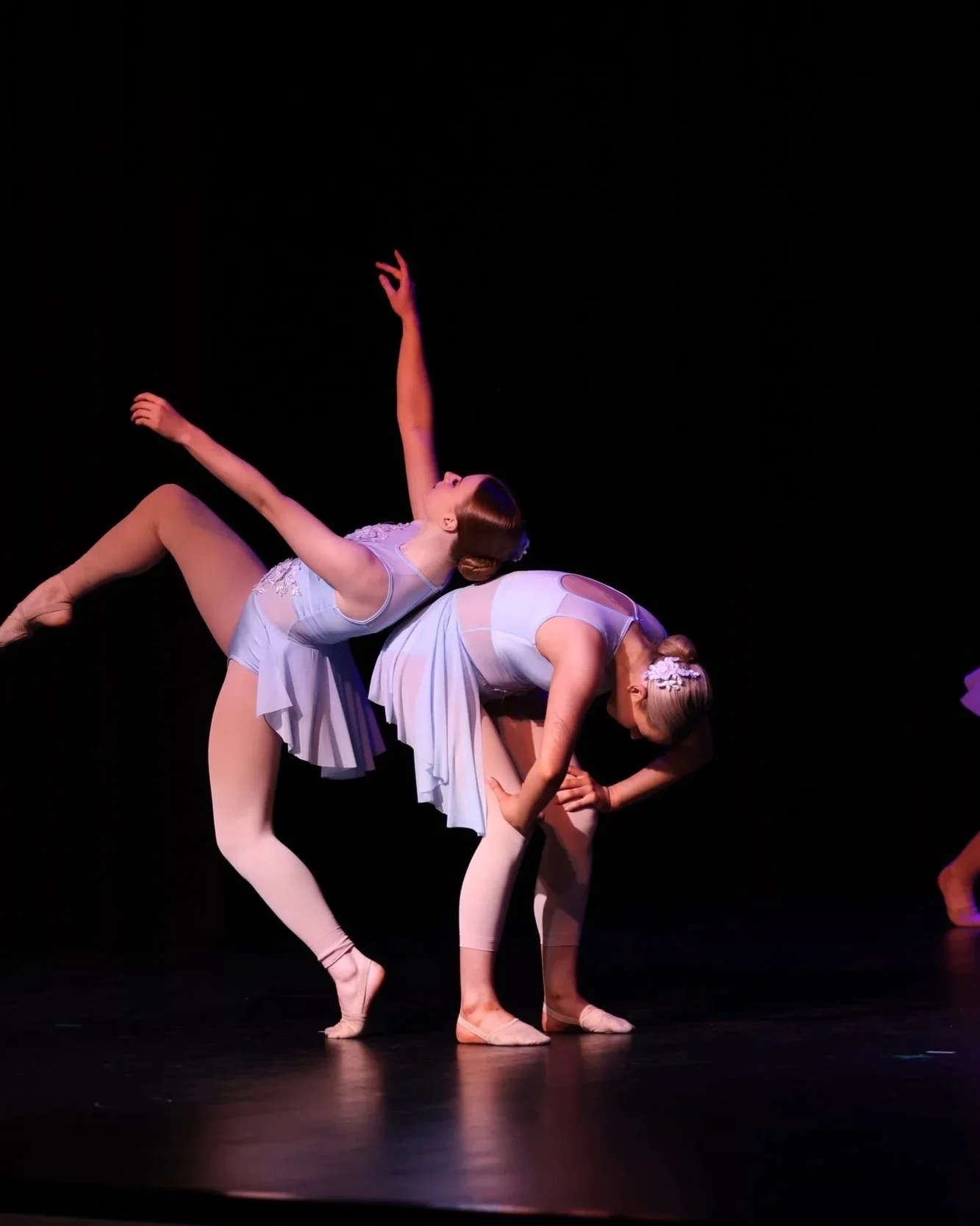 Two ballet dancers perform a pose on stage, with one dancer bending forward and supporting herself with her arms while the other dancer balances with an extended leg and arm, all dressed in light blue costumes and tights, against a dark background.
