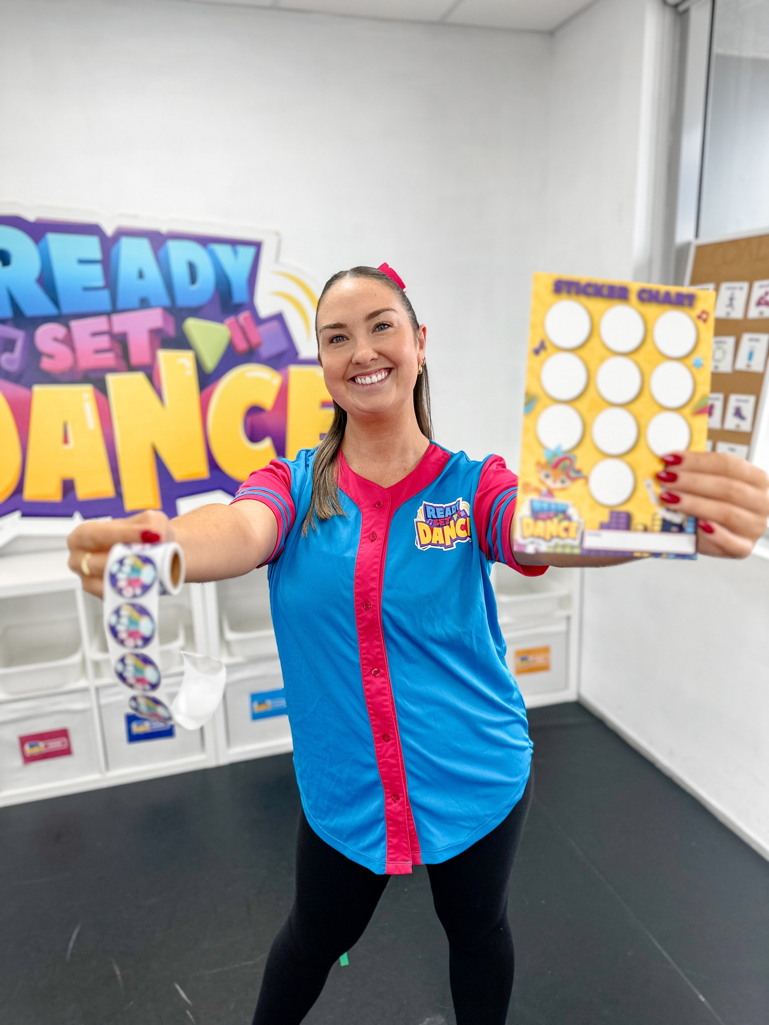 A woman smiling and holding a sticker chart and a roll of stickers, standing in a dance studio with a colorful sign in the background that reads 'Ready, Set, Dance'.