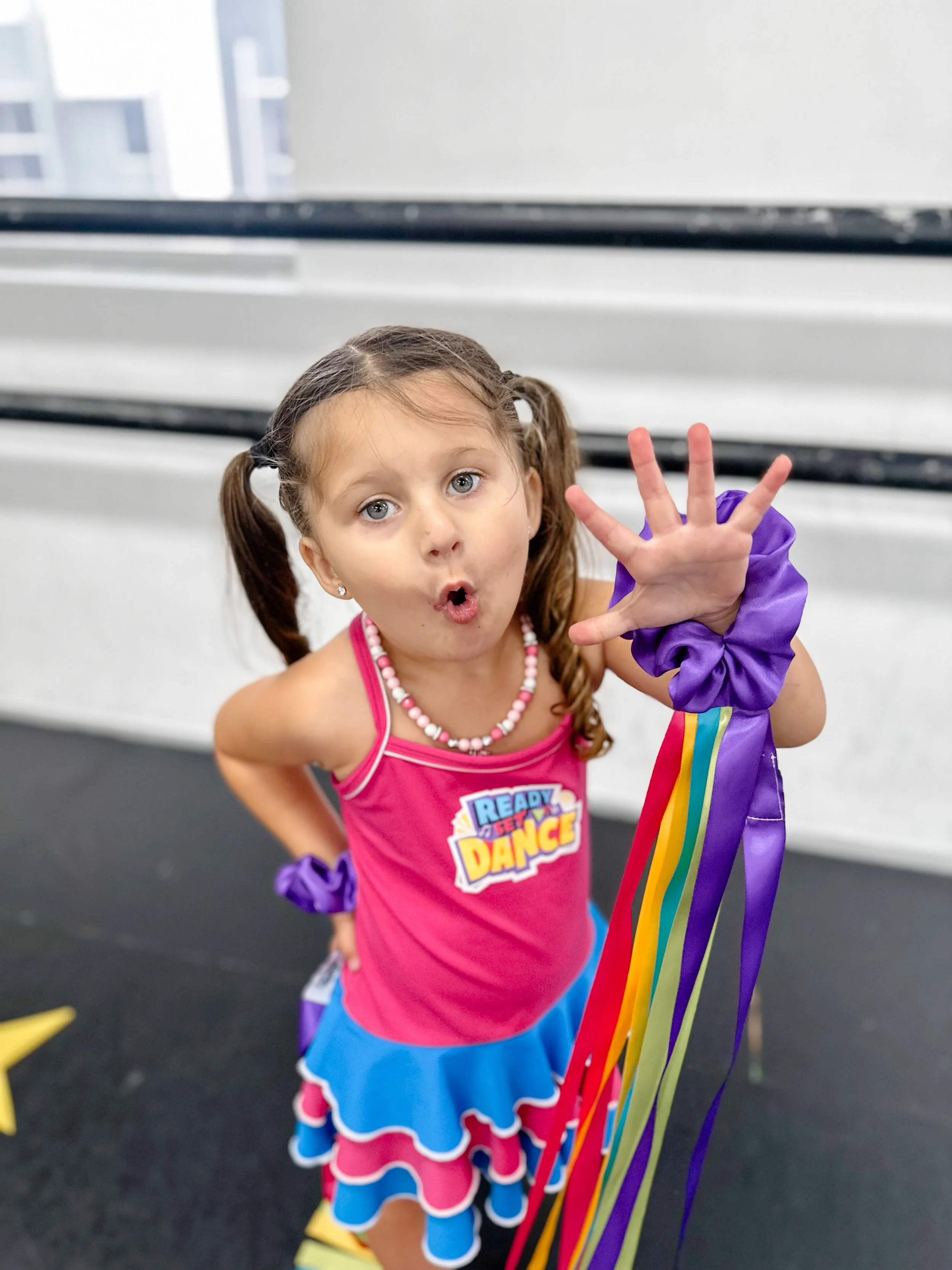 Young girl in a pink and blue dance costume holding colorful ribbons, making an expressive face.