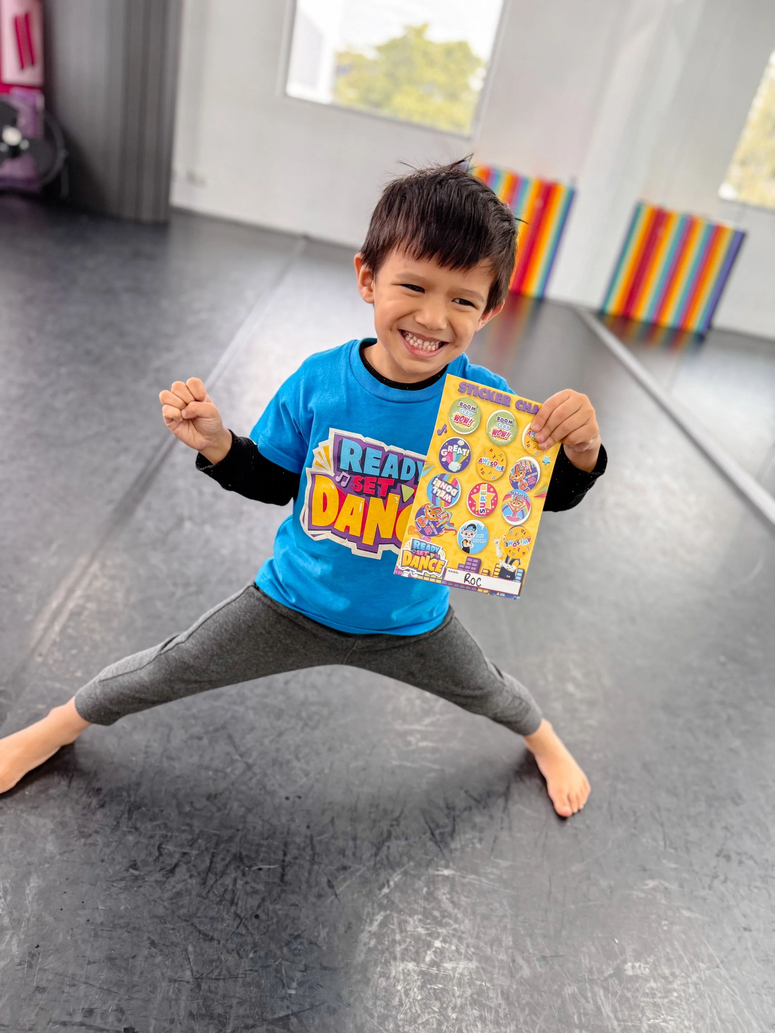 A young boy in a dance studio holding a sheet of colorful stickers while performing a dance move with a big smile.