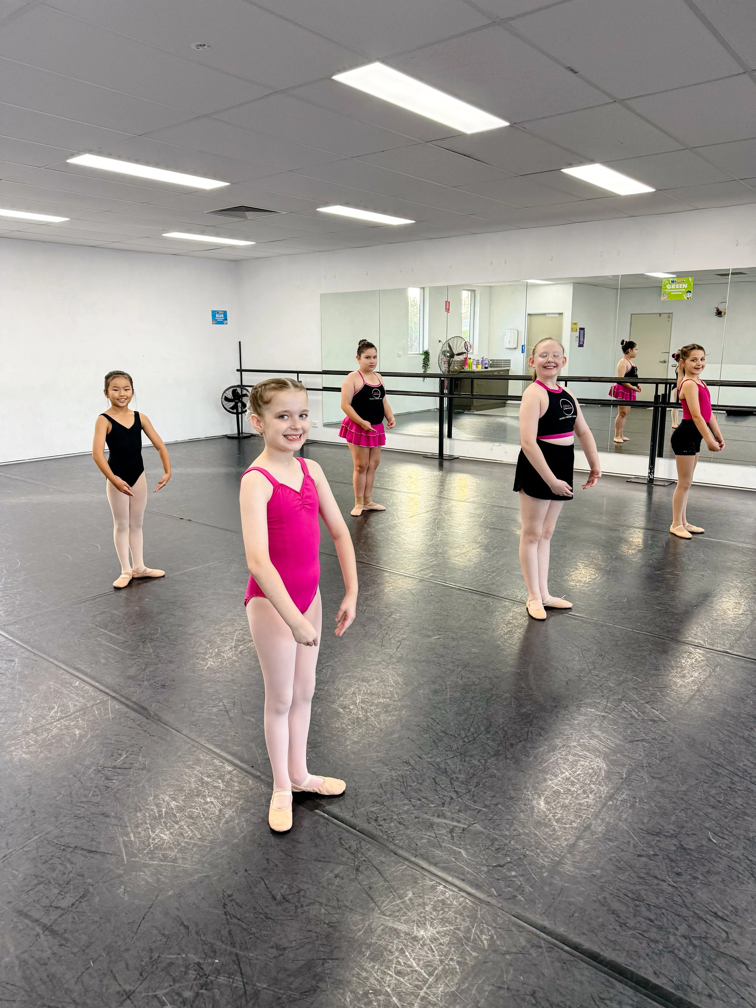 Young girls in ballet practice room wearing dance attire, standing at a ballet studio with mirrors, ballet barres, and a black floor.