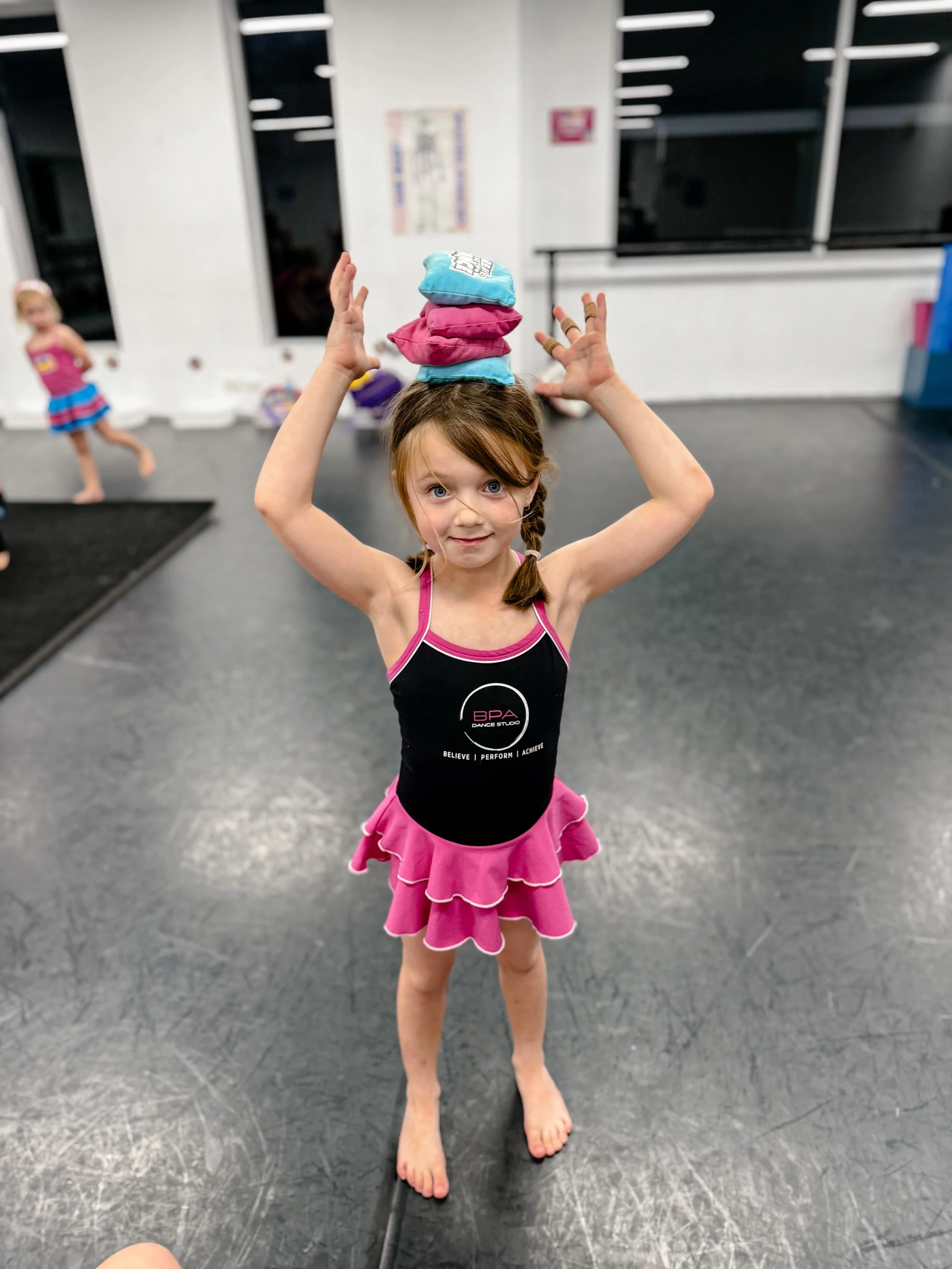 A young girl in a black and pink dance leotard and ruffled pink skirt balancing on her toes on the dance studio floor, with three colorful socks stacked on her head, inside a dance studio.