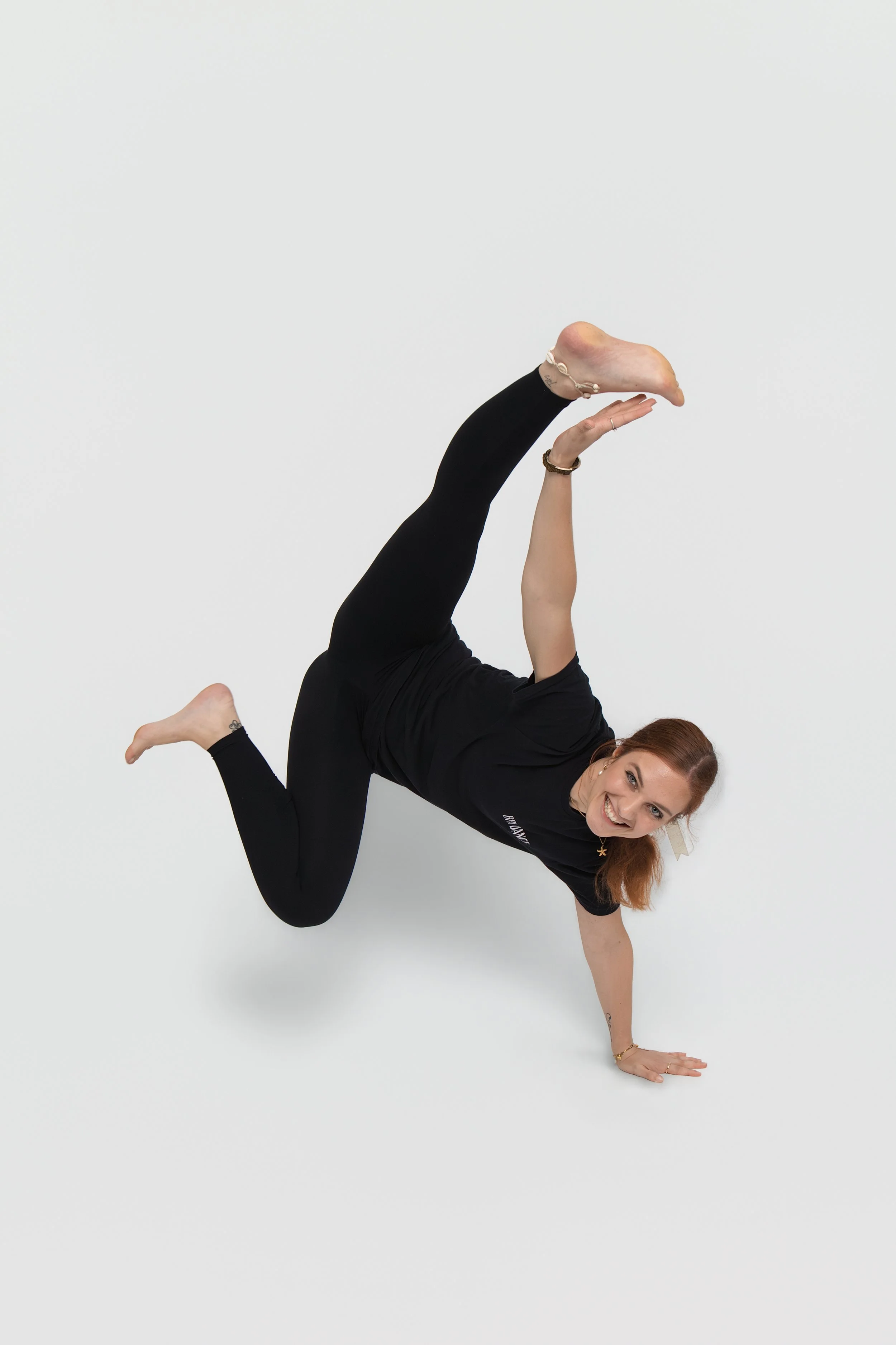 Woman with reddish-brown hair performing a one-handed handstand balanced on her left arm, smiling, wearing a black t-shirt and black leggings against a plain white background.