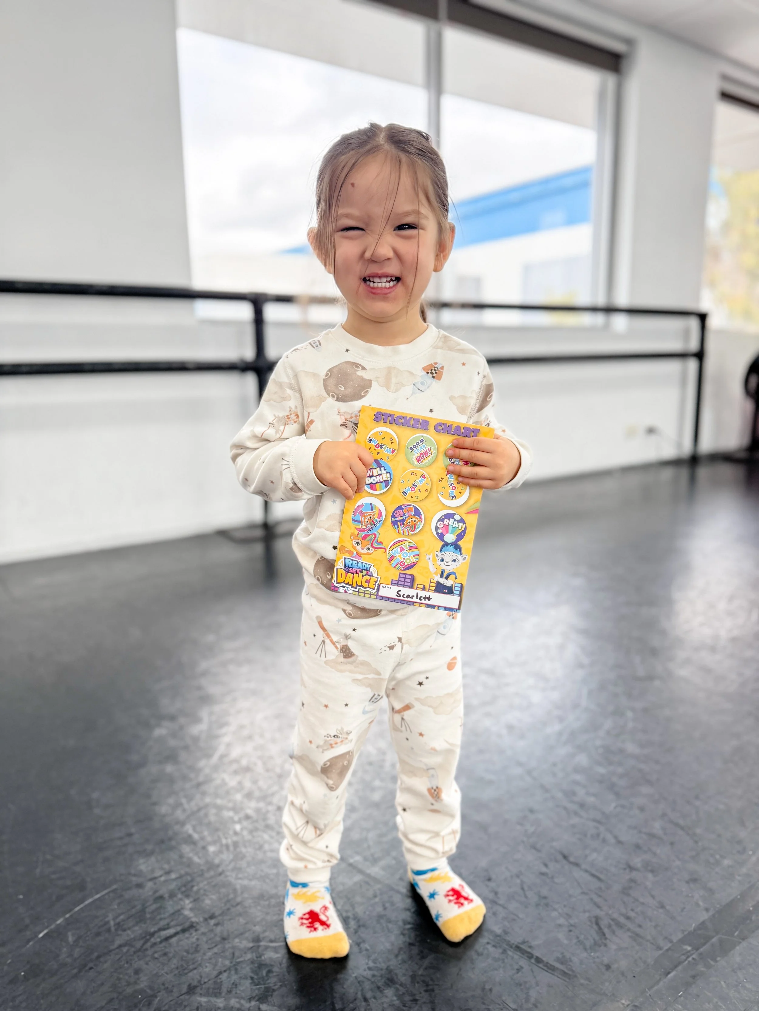 A young girl with a big smile, holding a sticker chart, standing indoors near large windows, wearing pajamas with a woodland animal print and colorful socks with dragons.