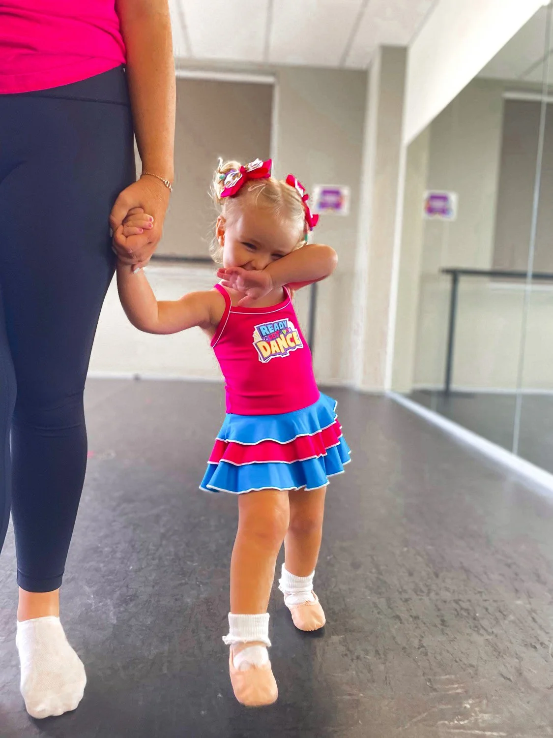 A young girl with blonde hair tied with pink and white bows, wearing a bright pink and blue cheerleading outfit with the words 'Ready to Dance' on her top, is smiling and covering her mouth with her arm while holding hands with an adult.