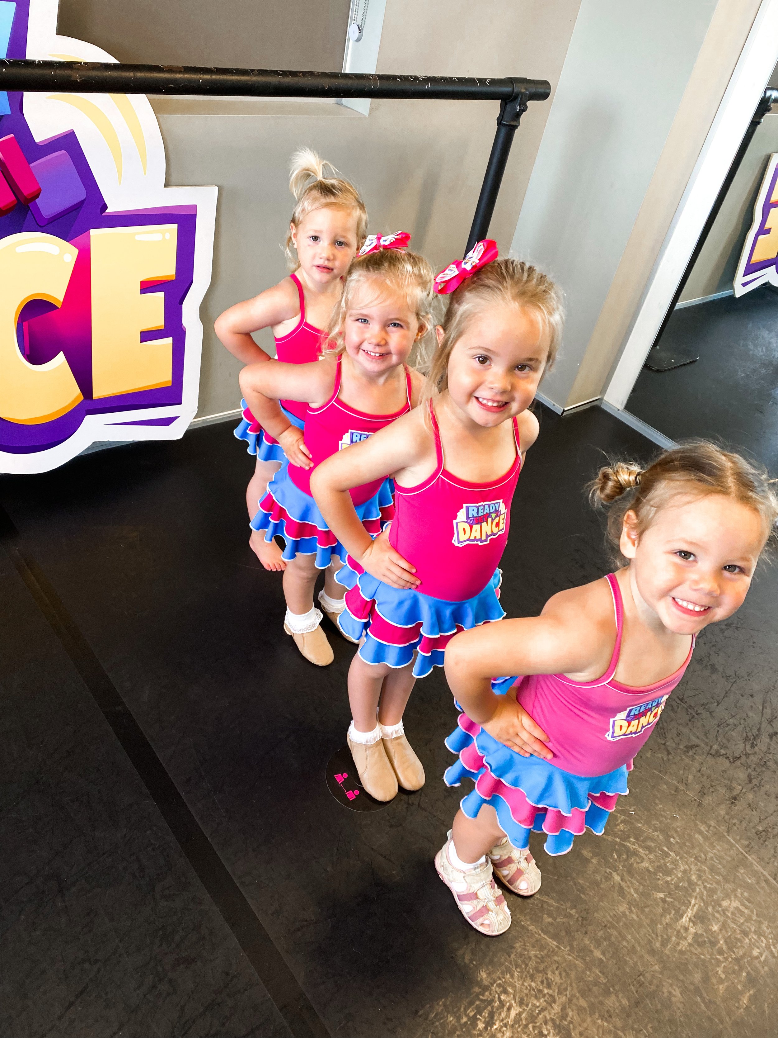 Four young girls in pink and blue cheerleading outfits standing in a line with hands on their hips, smiling at the camera, in a dance studio with a mirrored wall and a large colorful sign that says 'ICE' in the background.