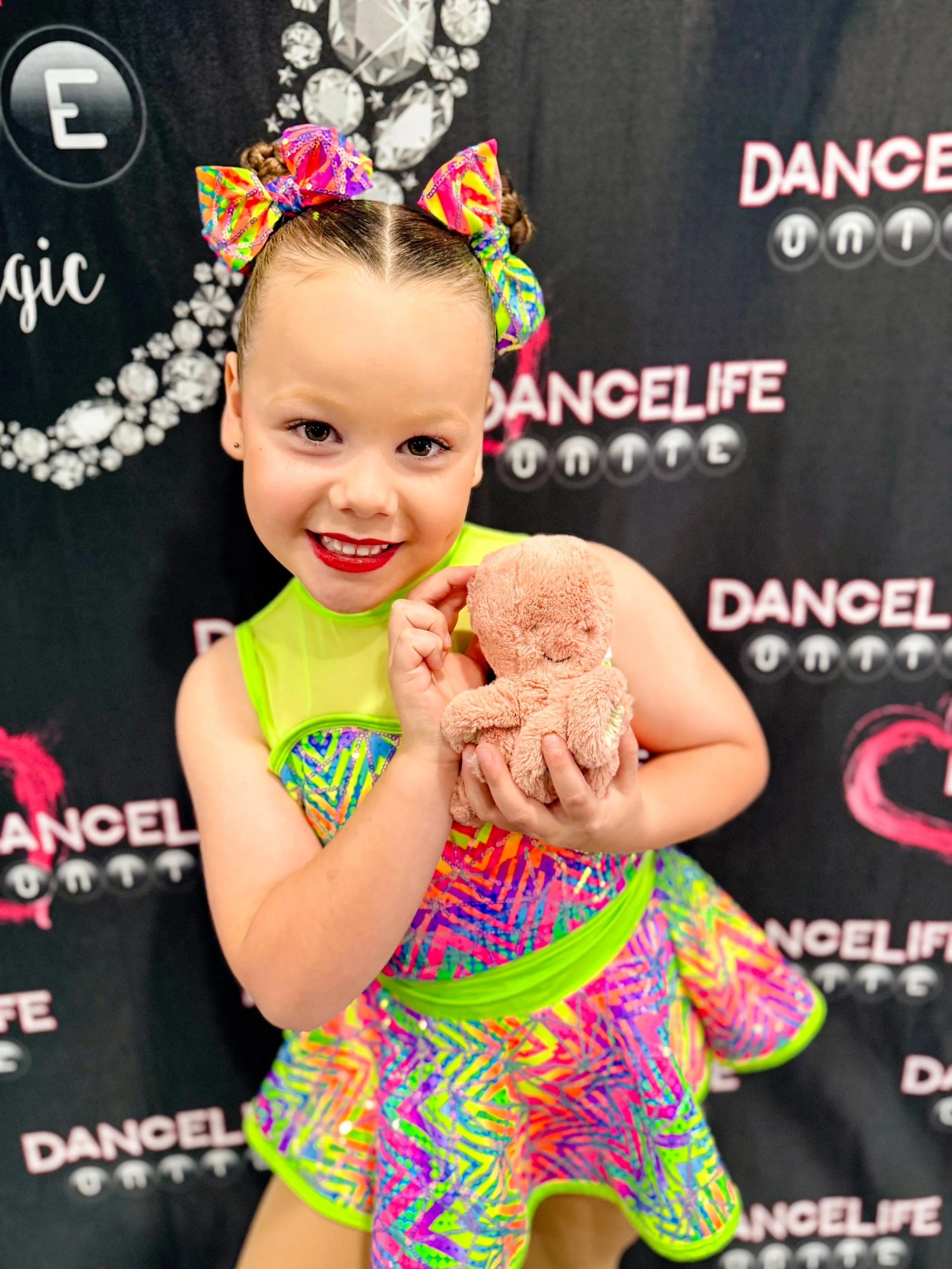 A young girl in a colorful dance costume with neon yellow accents and a matching hair bow, smiling and holding a small plush teddy bear. The background features a DanceLife logo and dance-themed decorations.