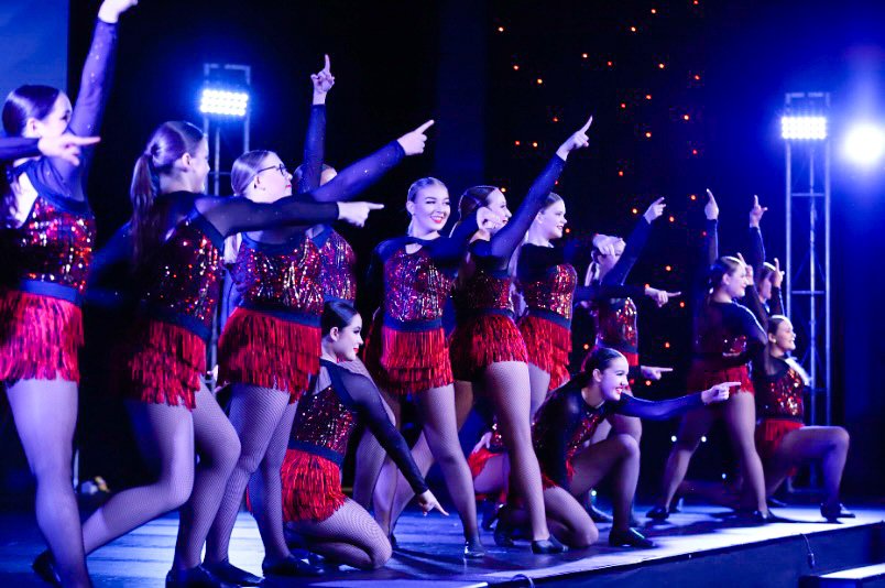 A group of young female dancers performing on stage, wearing matching red and black costumes with fringes, under stage lighting.