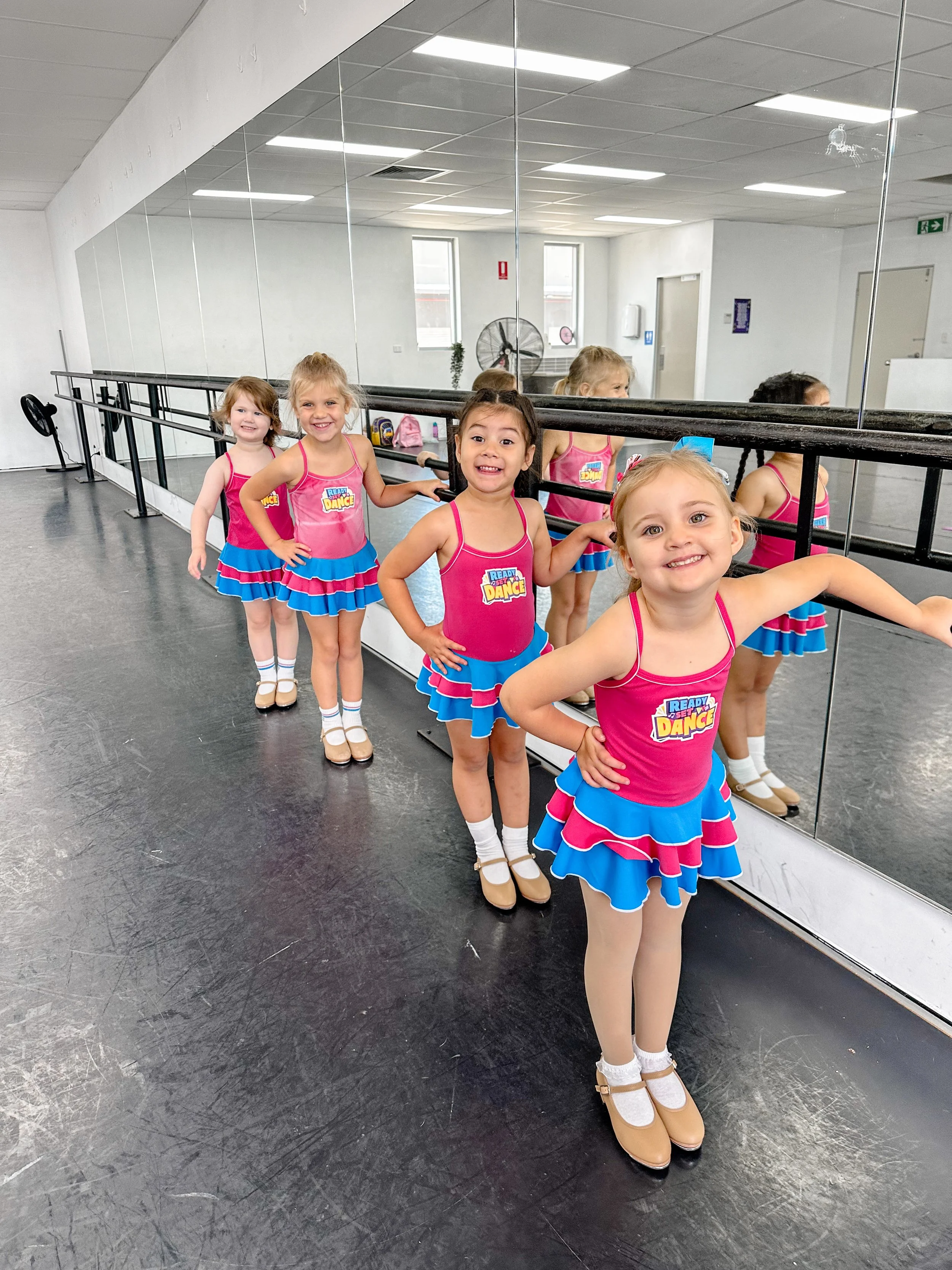 Four young girls in pink and blue dance costumes standing in line at a ballet studio, with their reflections visible in the mirror behind them.