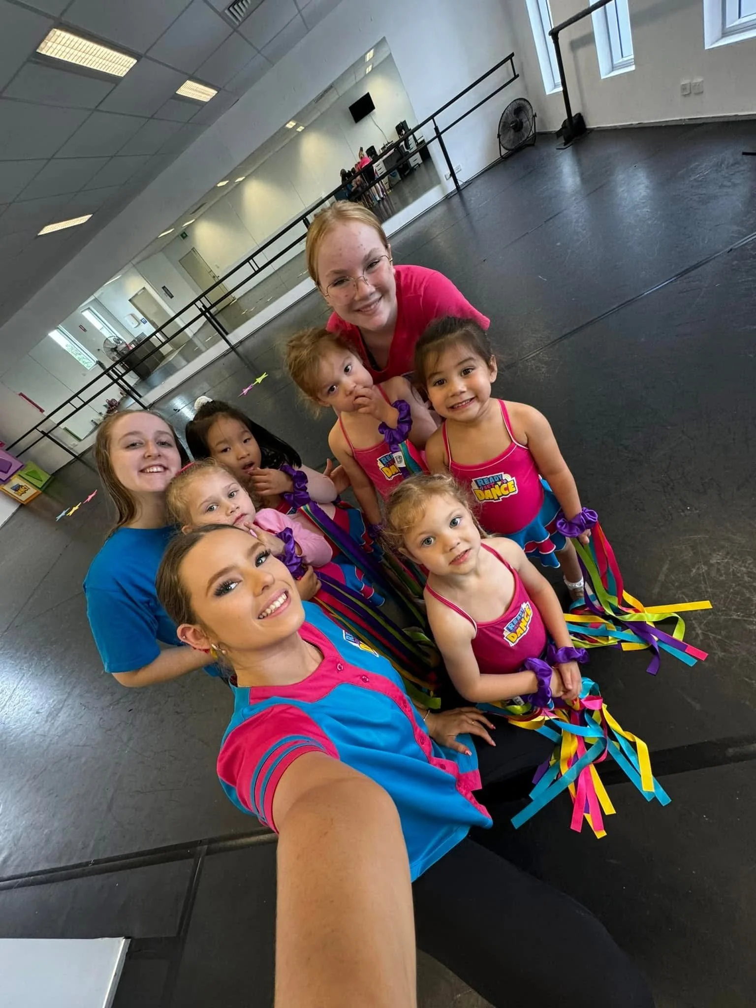 A woman taking a selfie with a group of young girls in a dance studio, all smiling and wearing colorful dance costumes with ribbons.
