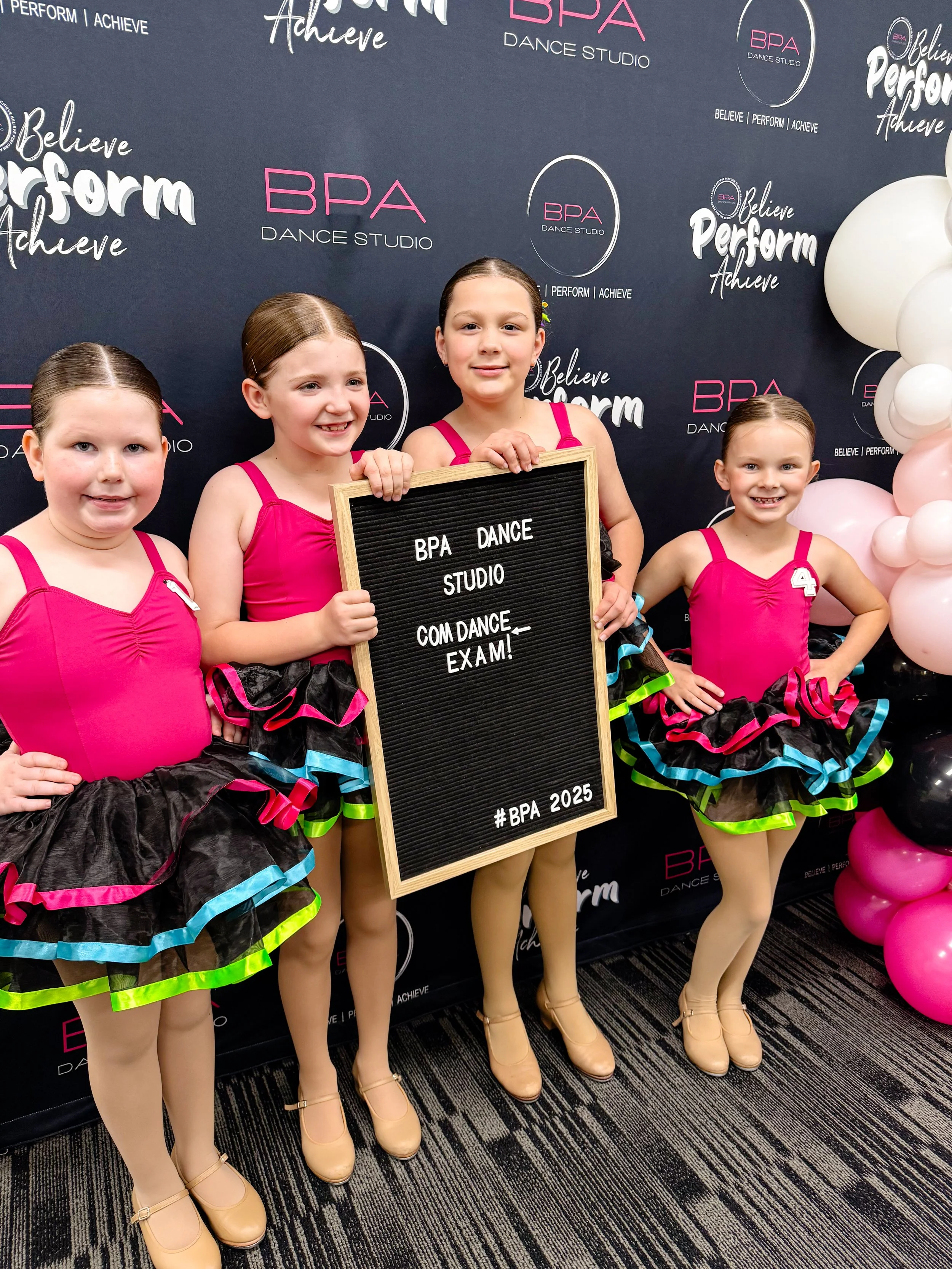 Four young girls in pink and black dance costumes with ruffled skirts, standing in front of a black backdrop with pink and white text and balloons, holding a sign that reads, "BPA Dance Studio, COM DANCE, EXAM! #BPA 2025."