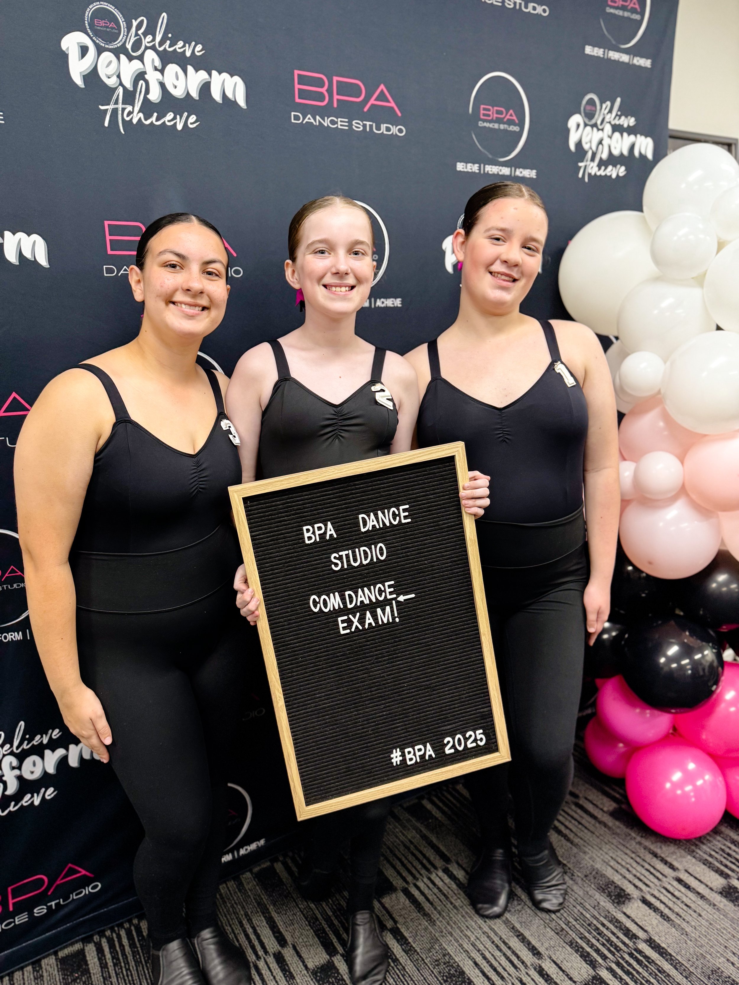 Three young women in black dance costumes standing together indoors at a dance studio event, holding a black message board that reads 'BPA Dance Studio Com Dance Exam! #BPA 2025', with thoughts of balloons to their right, and a backdrop with dance st