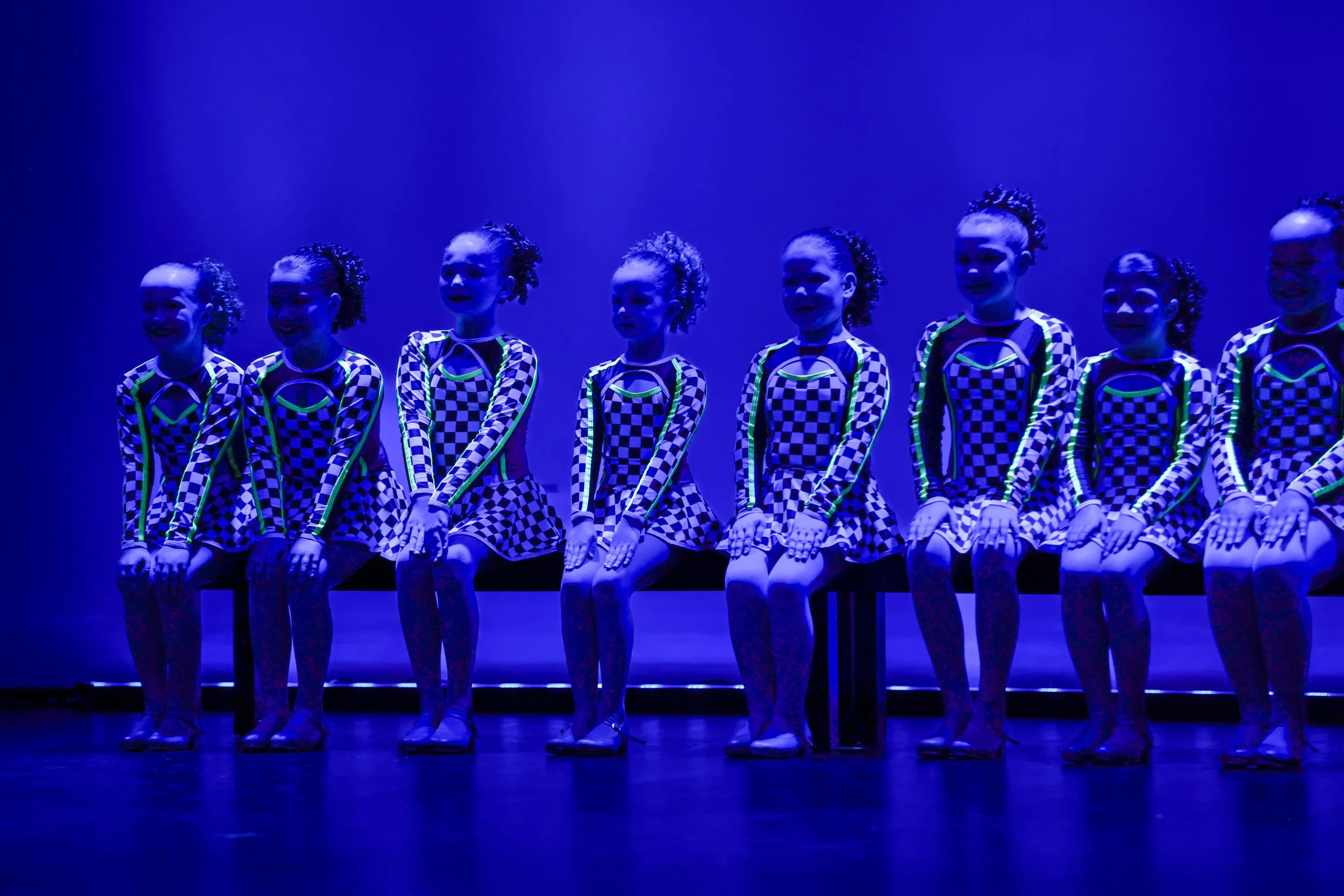 Eight young girls in matching checkered costumes sitting on a bench under blue stage lighting during a performance.