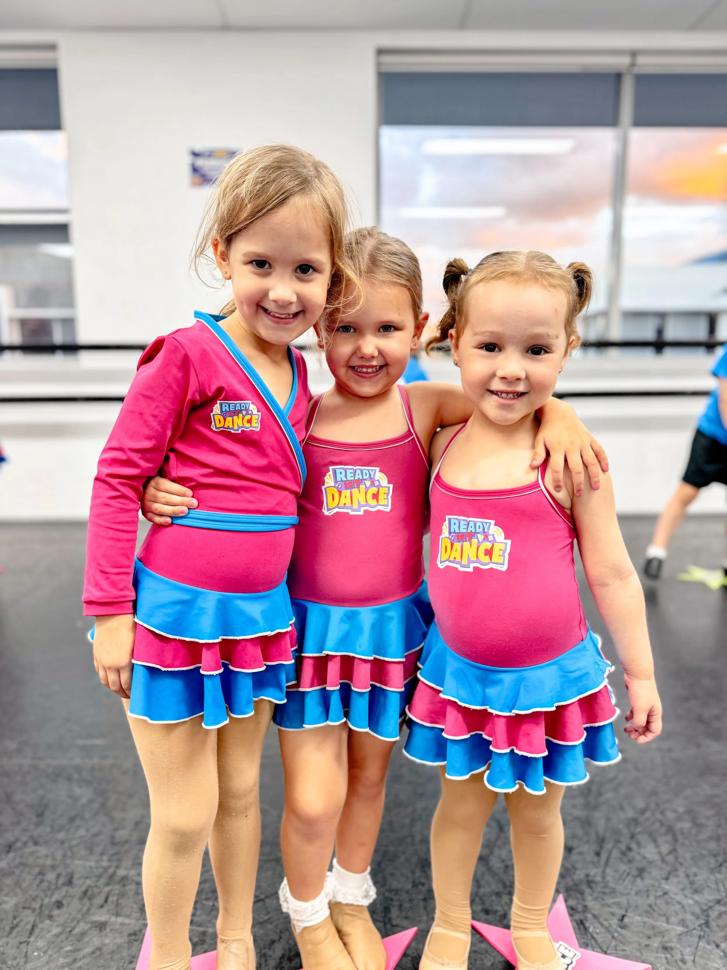Three young girls in pink and blue dance costumes smiling and posing together in a dance studio.