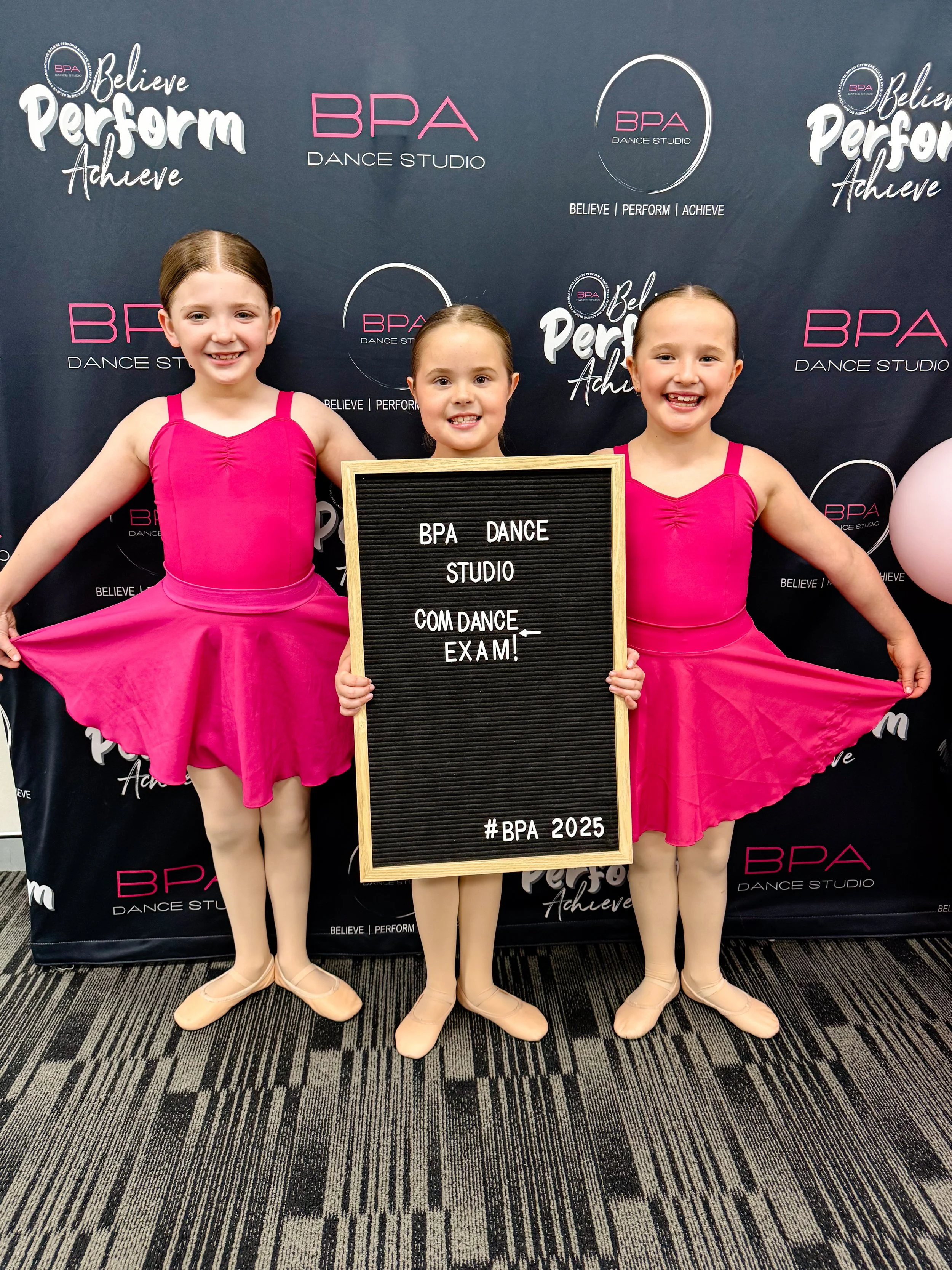 Three young girls in pink ballet costumes standing in front of a BPA Dance Studio backdrop, holding a sign that says they are ready for a dance exam.