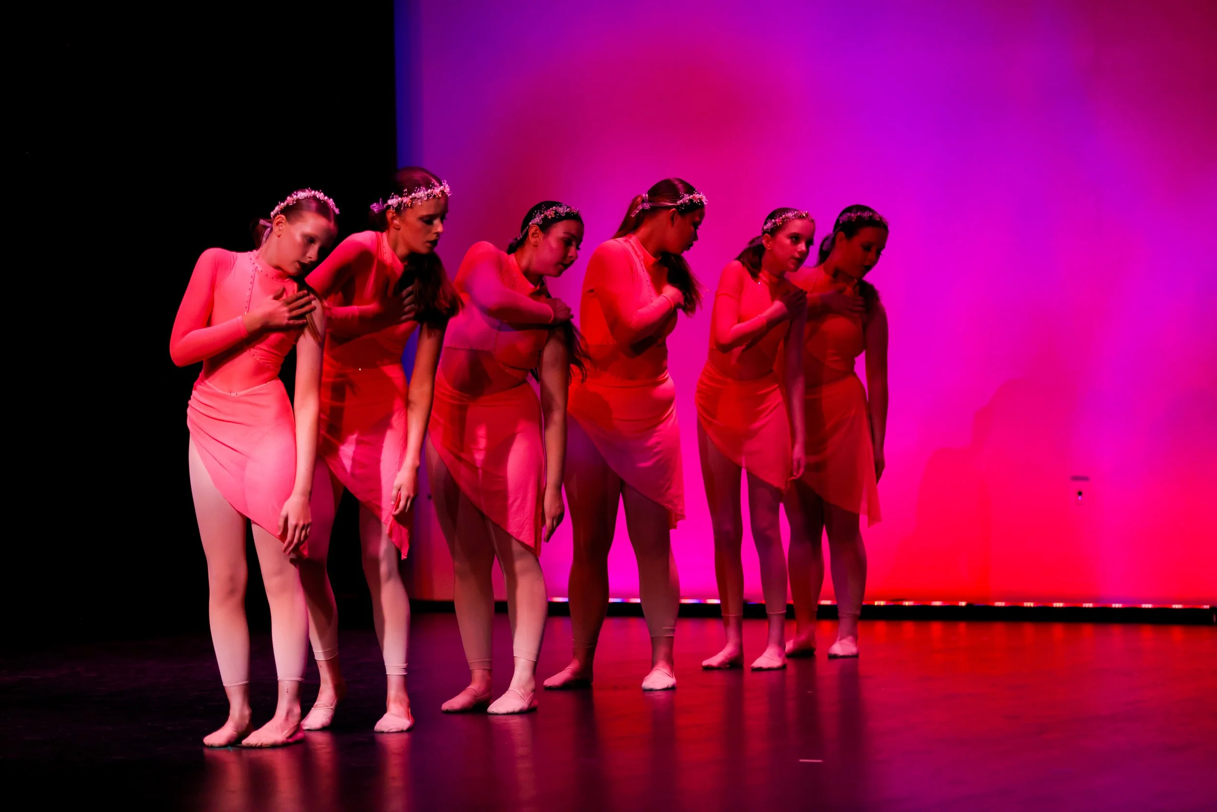 Six ballet dancers in pink dresses standing on stage with pink lighting, placing their right hands on their chests in a solemn pose.