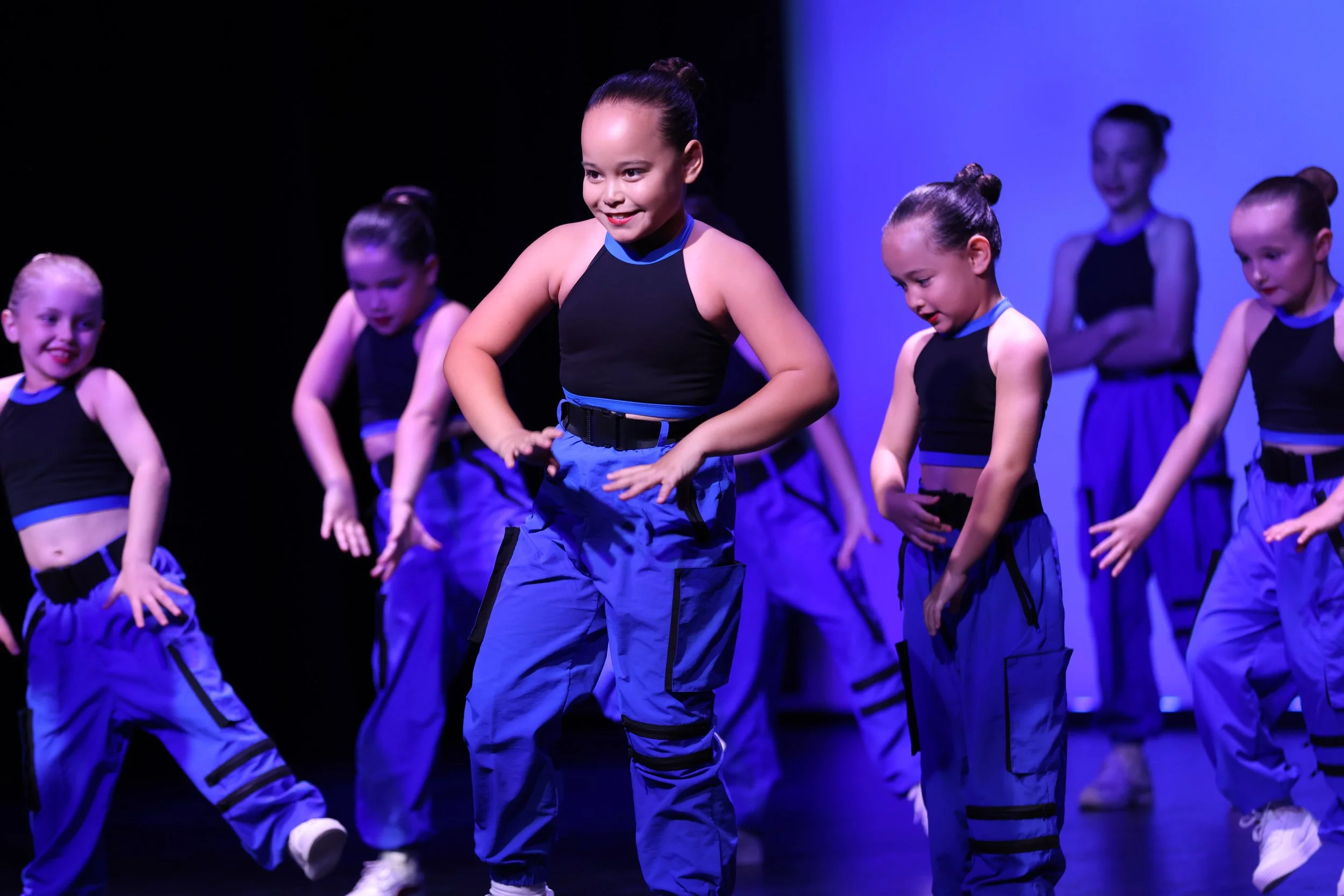 Young girls performing a dance routine on stage in matching black and blue outfits and baggy pants.