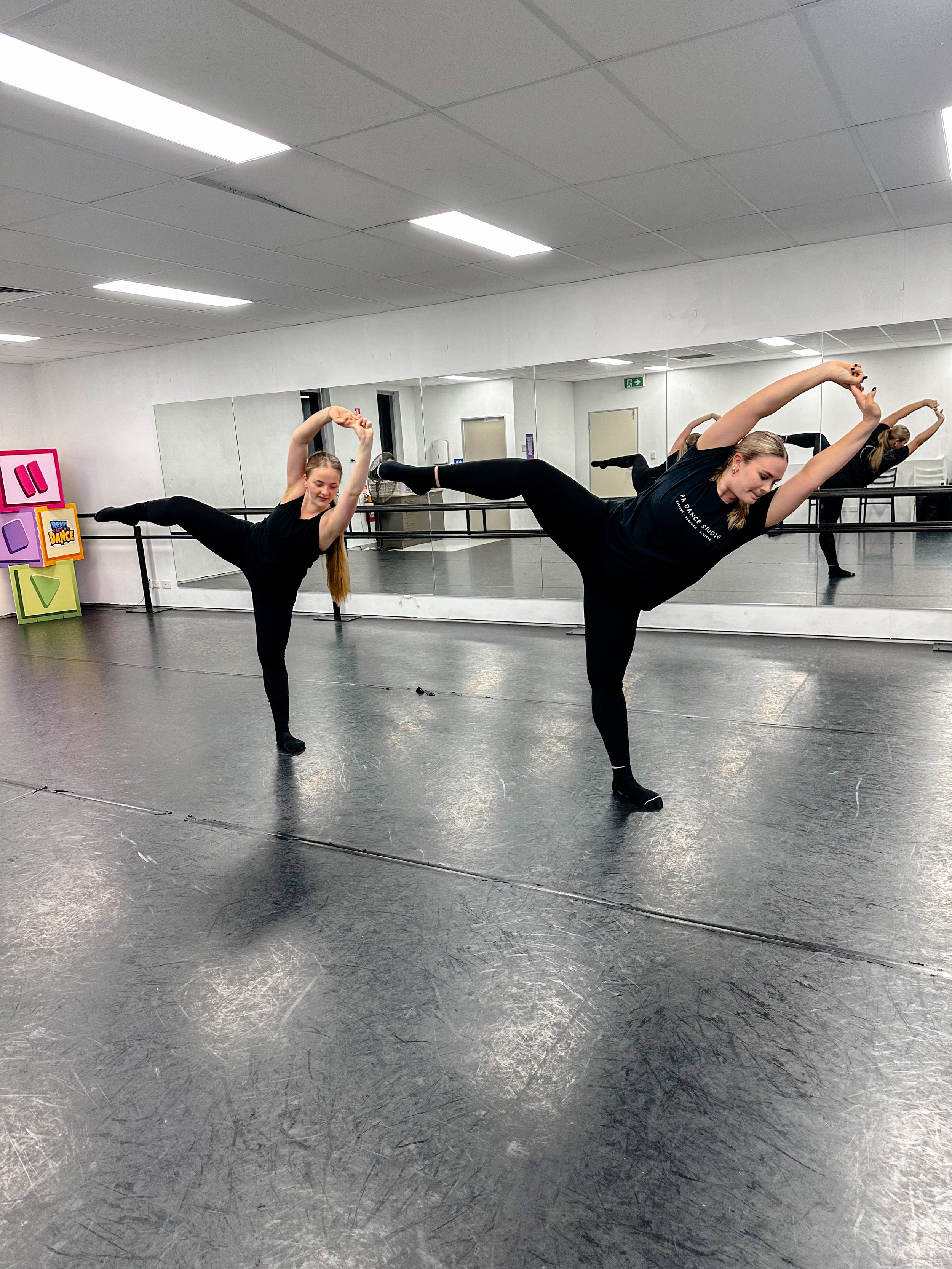 Two ballet dancers practicing in a dance studio, balancing on one leg with arms raised overhead and the other leg extended to the side, against a wall with a large mirror.