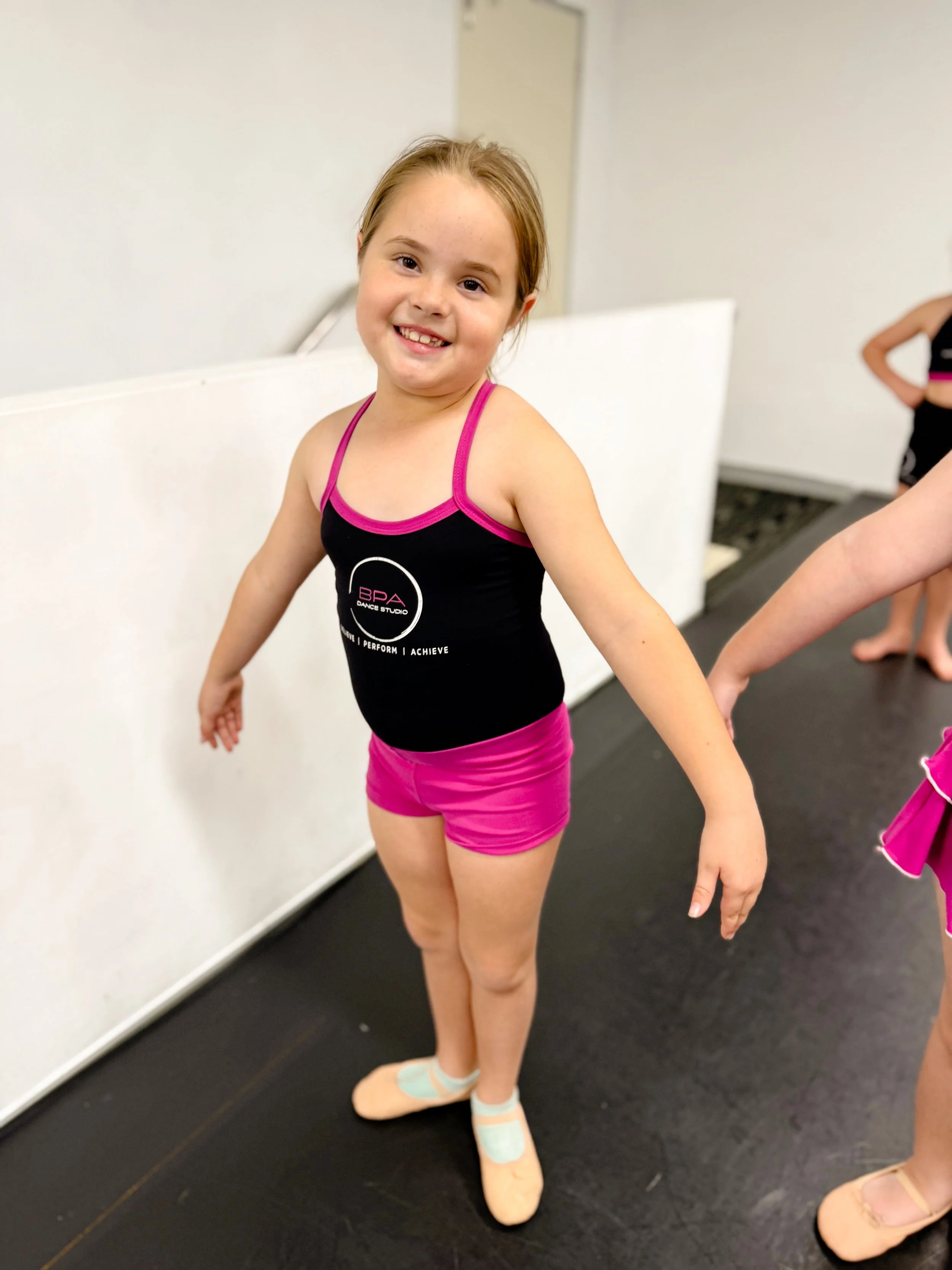 Young girl in ballet attire smiling at the camera, with dancers in the background at a dance studio.