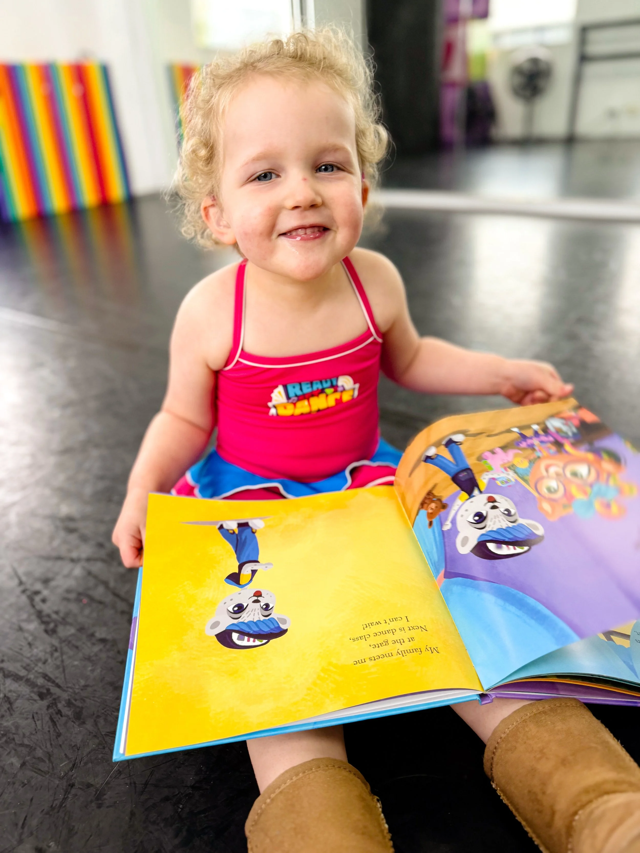 A young girl with curly blonde hair, wearing a pink tank top with the words 'Ready to Dance,' sitting on the floor and smiling while looking at a colorful children's book with animated characters.