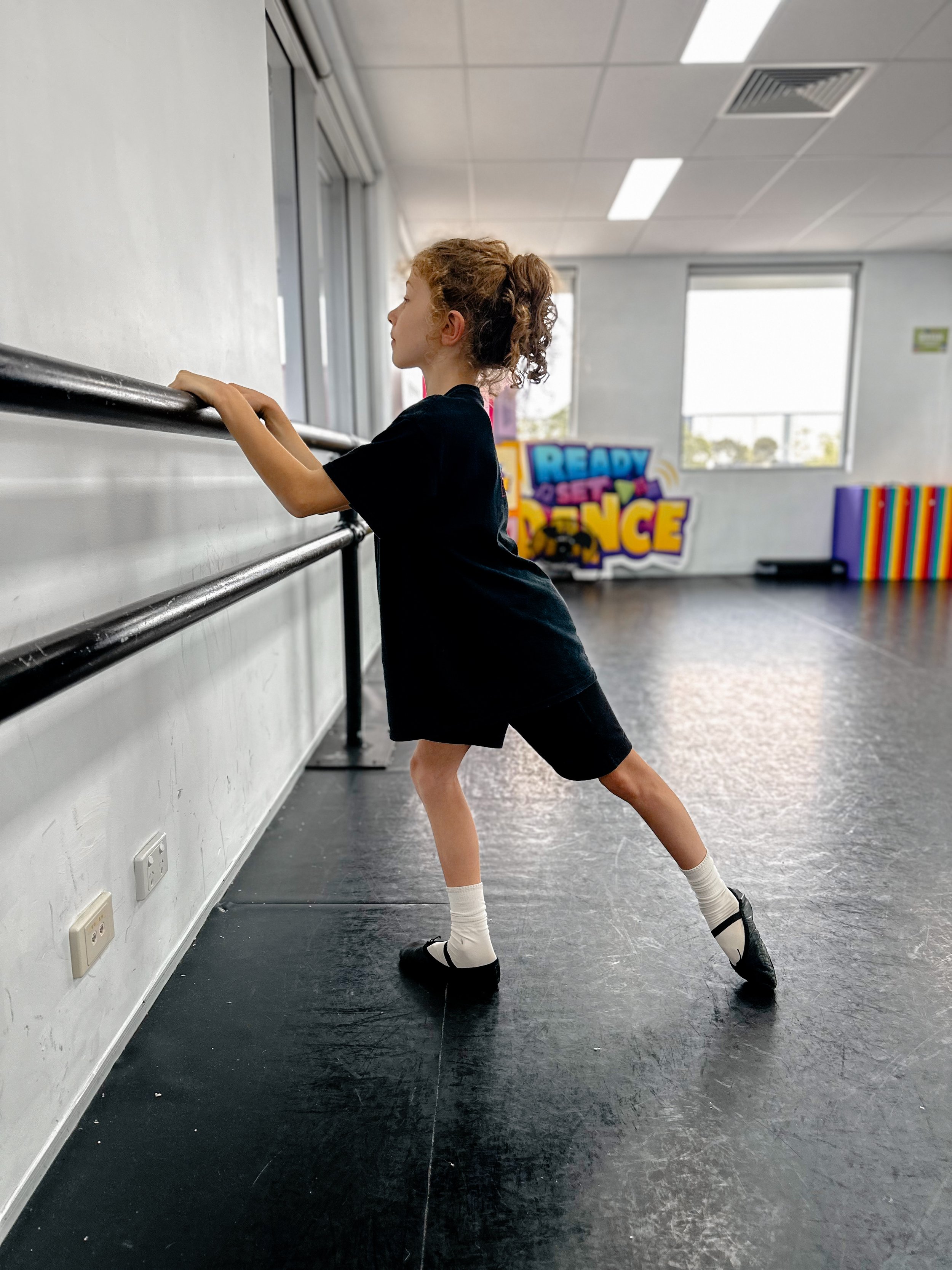 Young girl in black dance uniform stretching at a ballet barre in a dance studio with large windows and colorful workout equipment in the background.