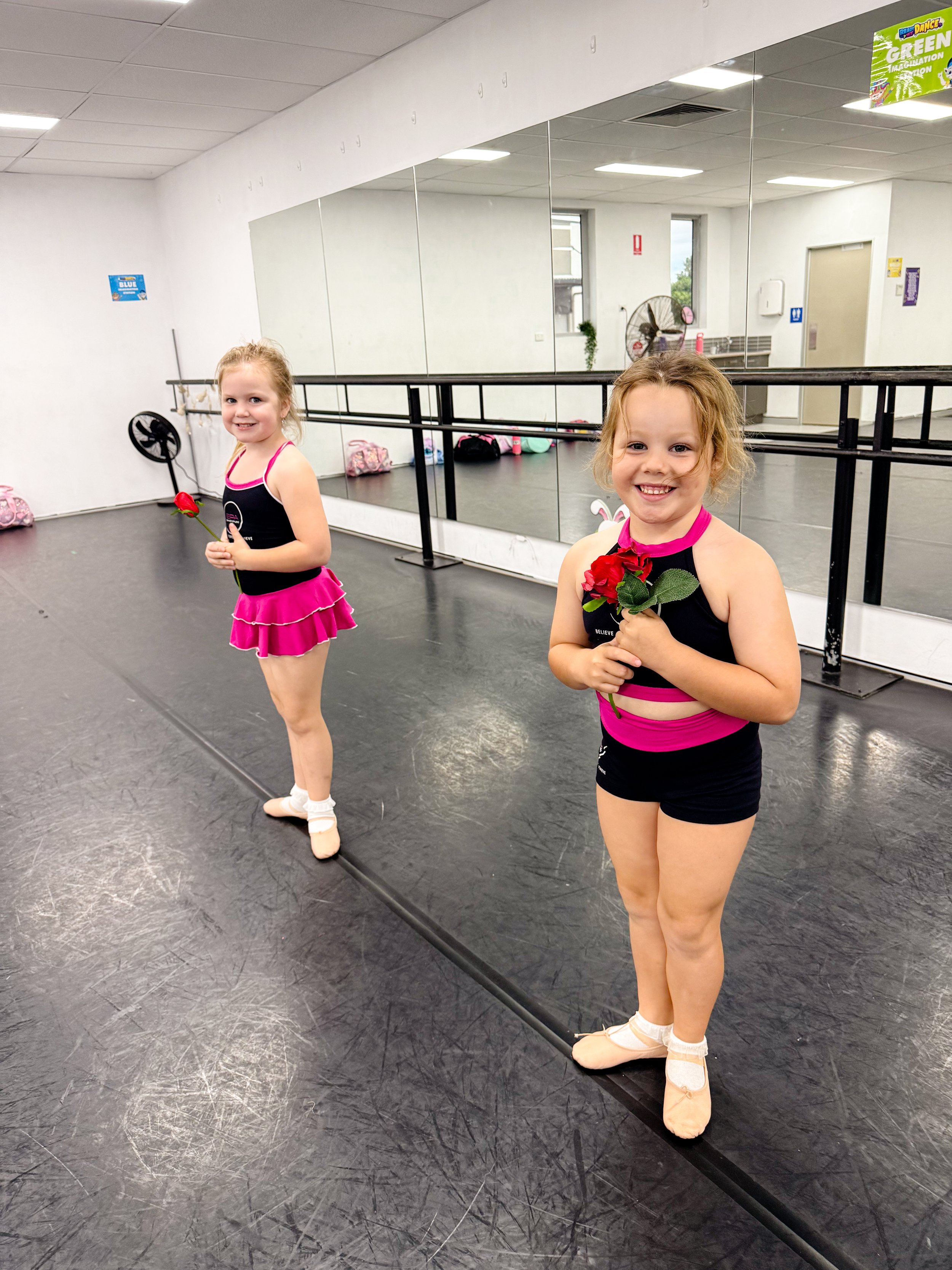 Two young girls in ballet costumes holding roses in a dance studio, smiling at the camera.