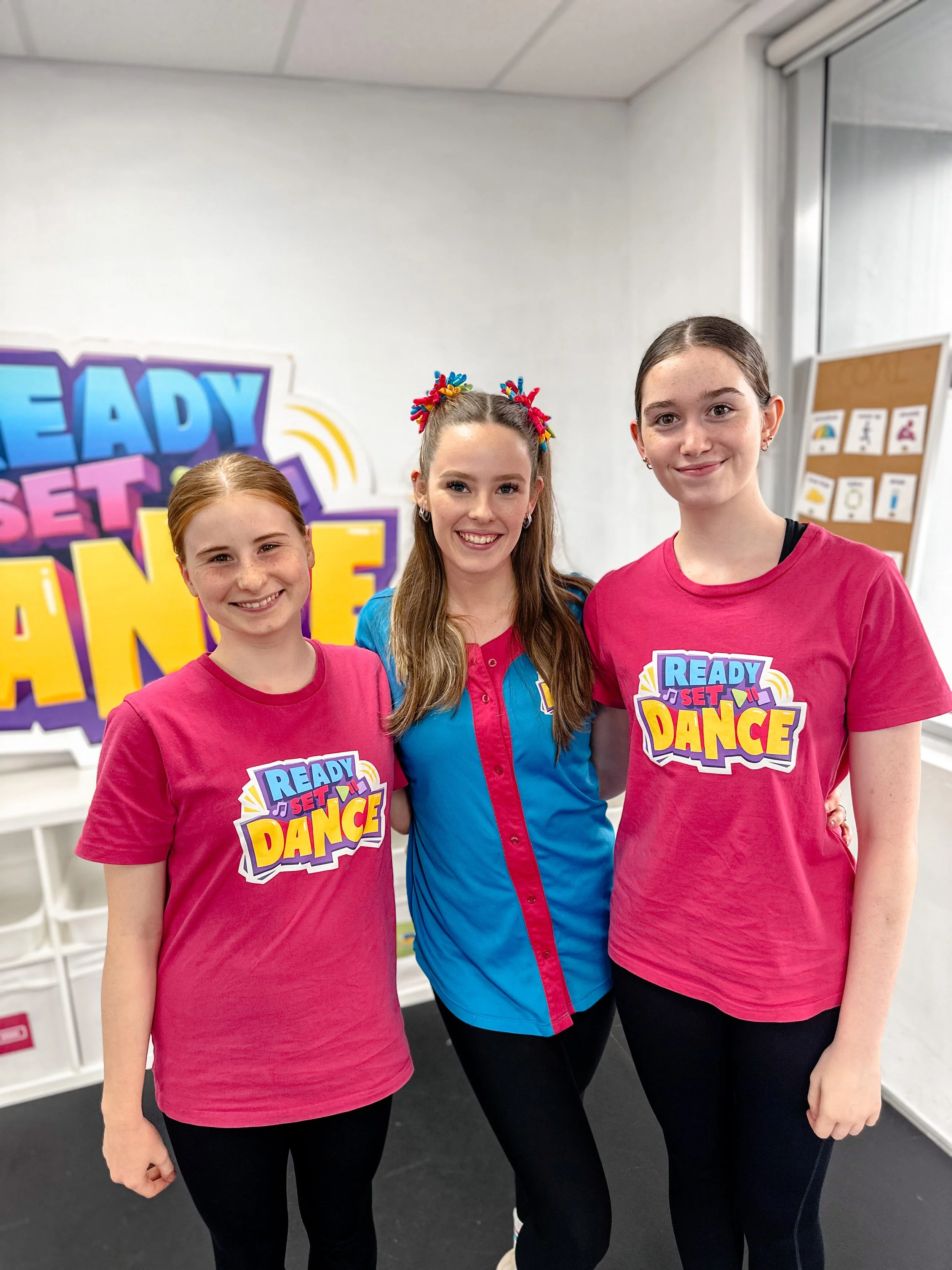 Three young girls smiling and posing together indoors, with colorful 'Ready Set Dance' signage in the background. Two girls are wearing pink t-shirts with the same logo, and one girl is wearing a blue and pink shirt.