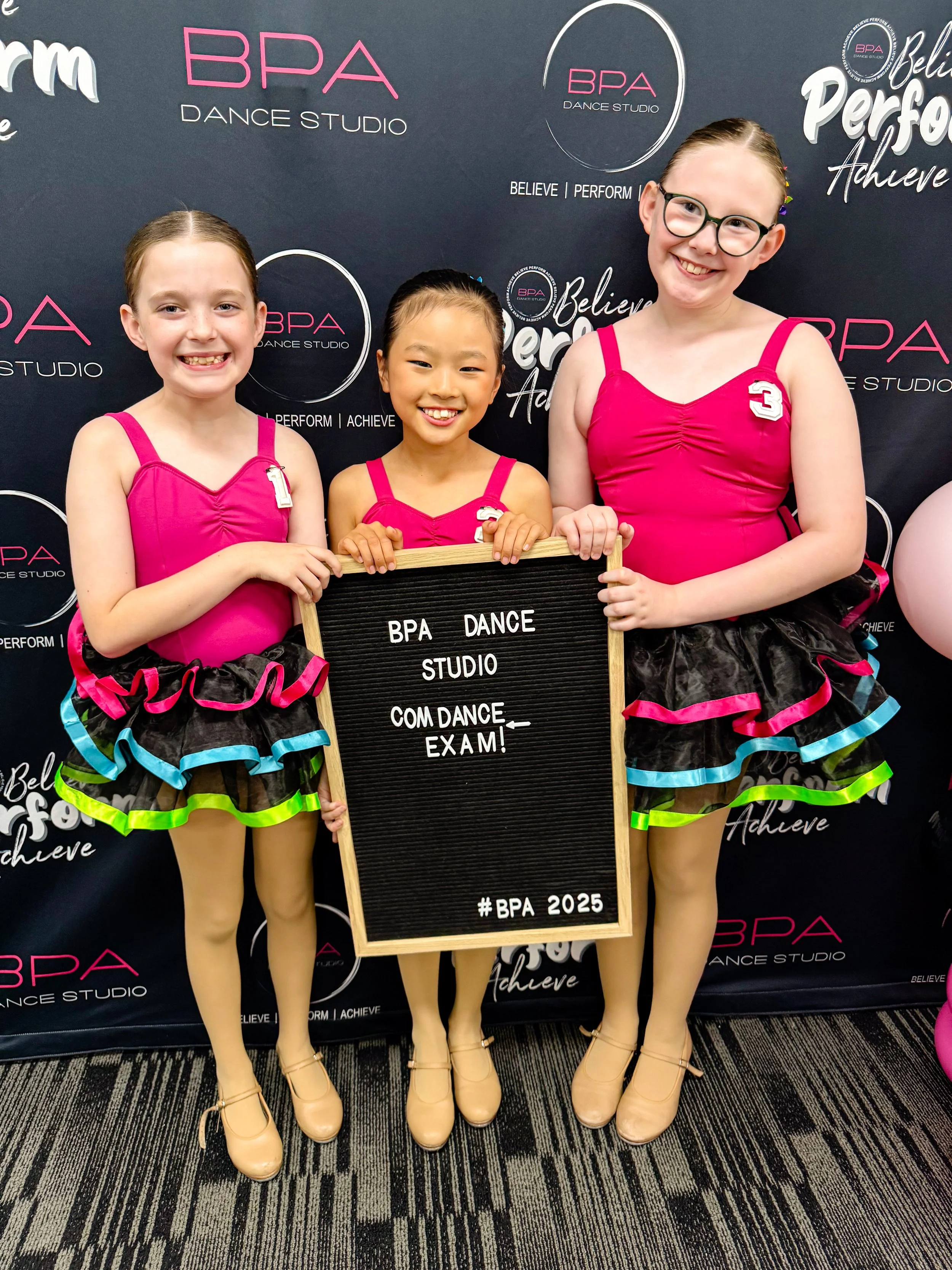 Three young girls in colorful dance costumes and beige dance shoes standing in front of a black backdrop with the BPA Dance Studio logo and slogans. They are smiling and holding a blackboard sign that reads 'BPA Dance Studio Commencing Exam!'