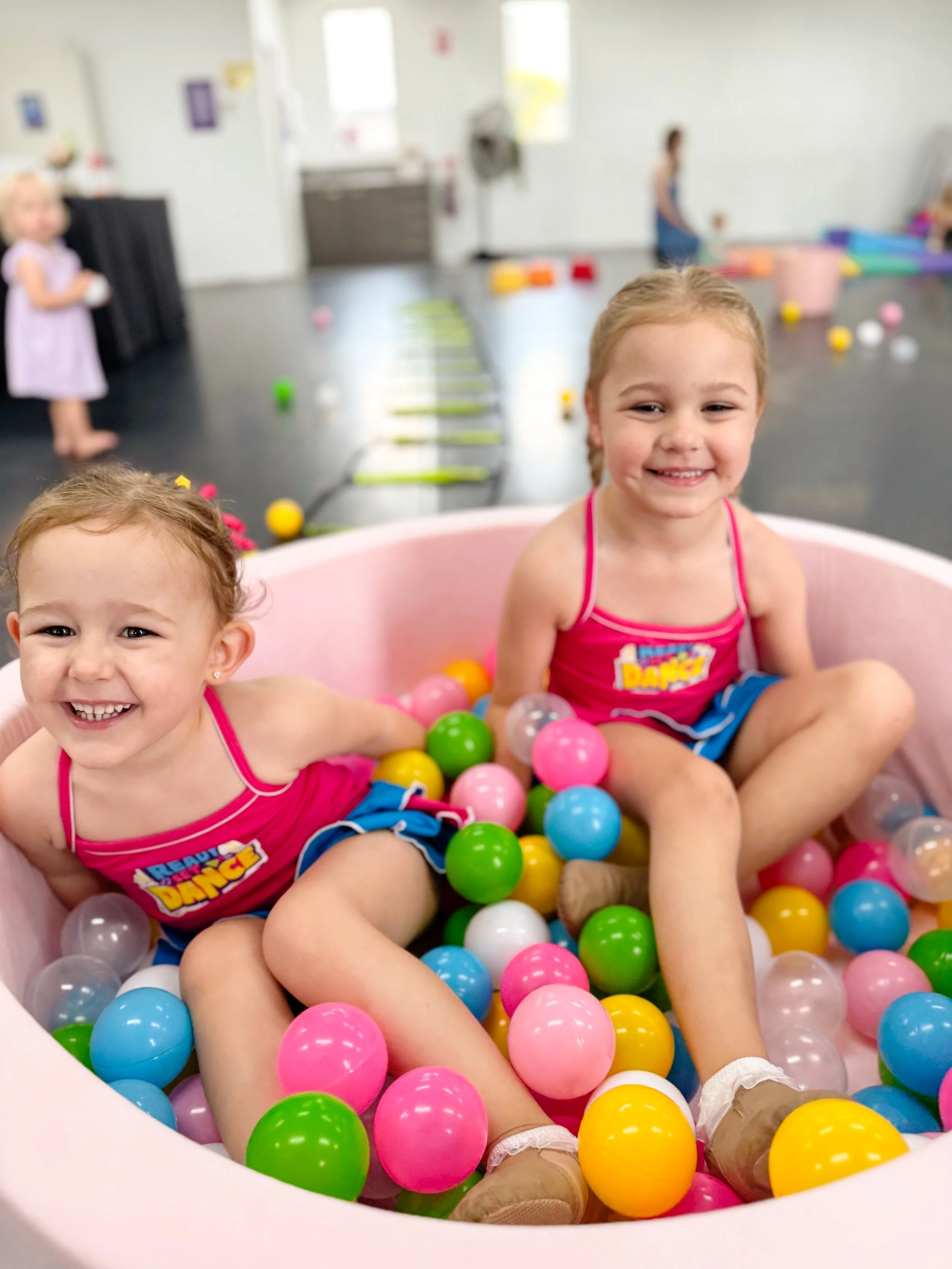 Two young girls smiling and sitting in a ball pit of colorful plastic balls in a playroom