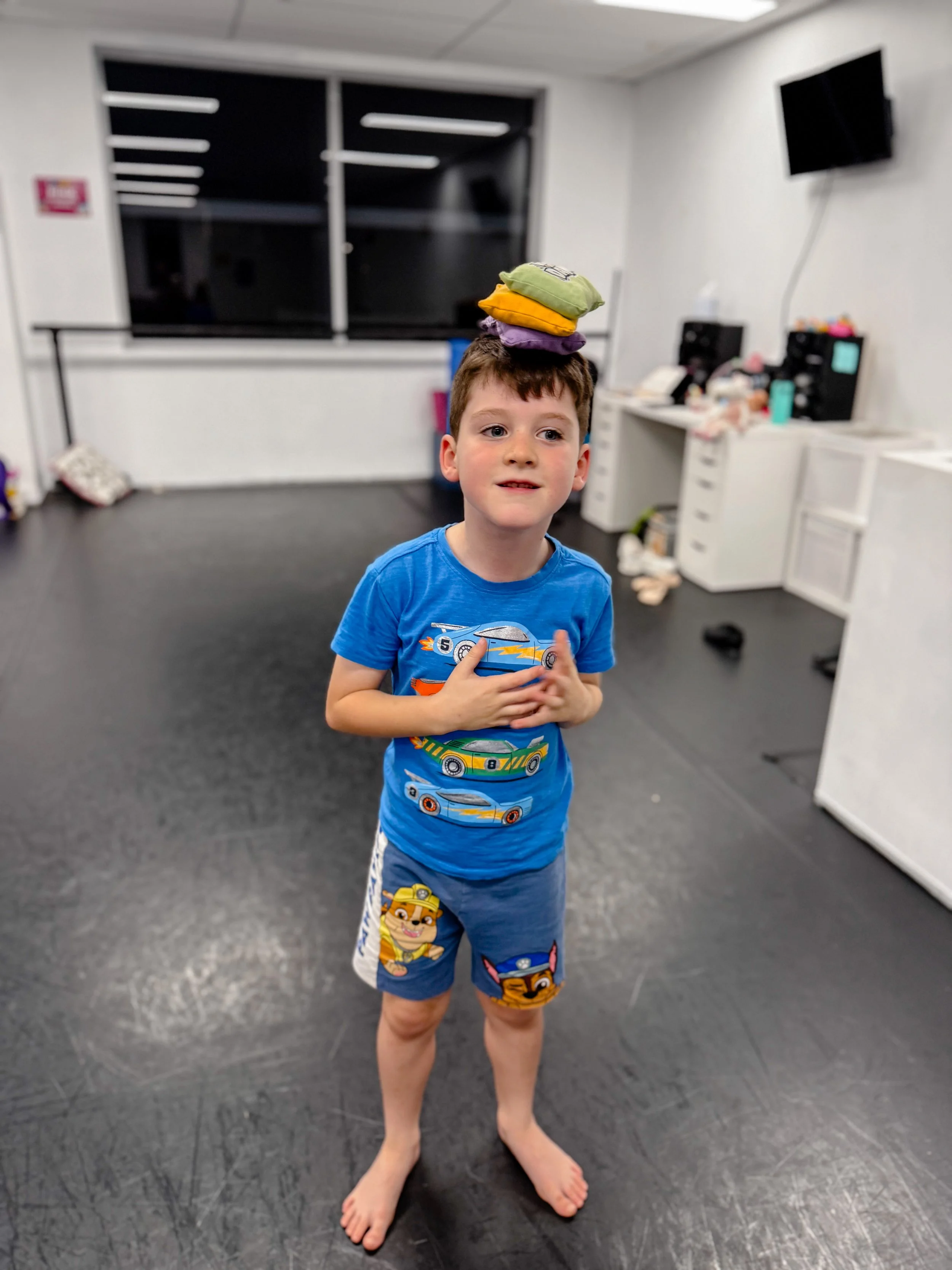 Boy standing indoors with a stack of colorful clothes on his head, wearing a blue car-themed t-shirt and shorts, with a playful expression.