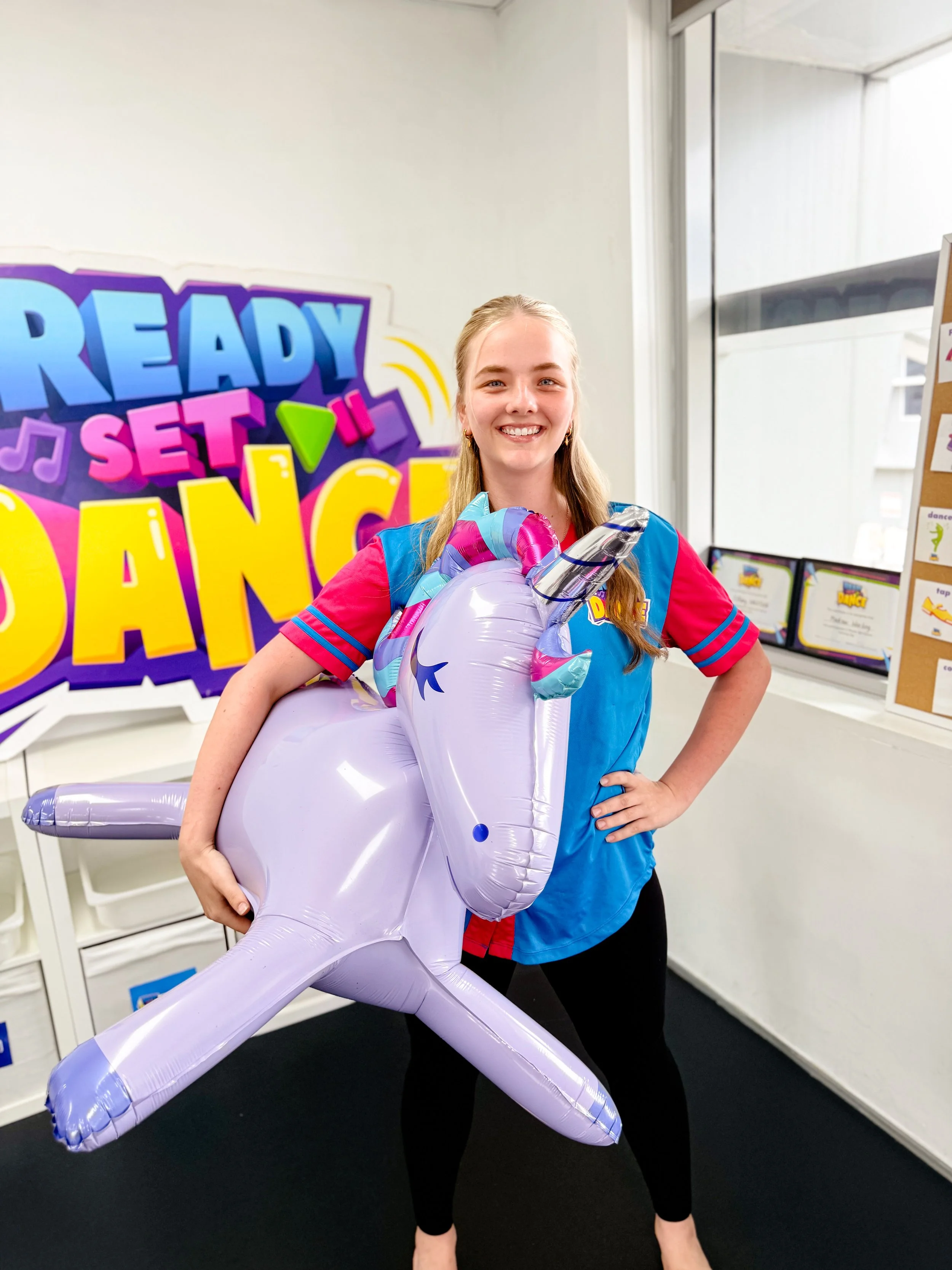 Young woman holding a large, purple unicorn-shaped balloon, smiling, standing in a room with colorful signage that reads "Ready Set Dance."