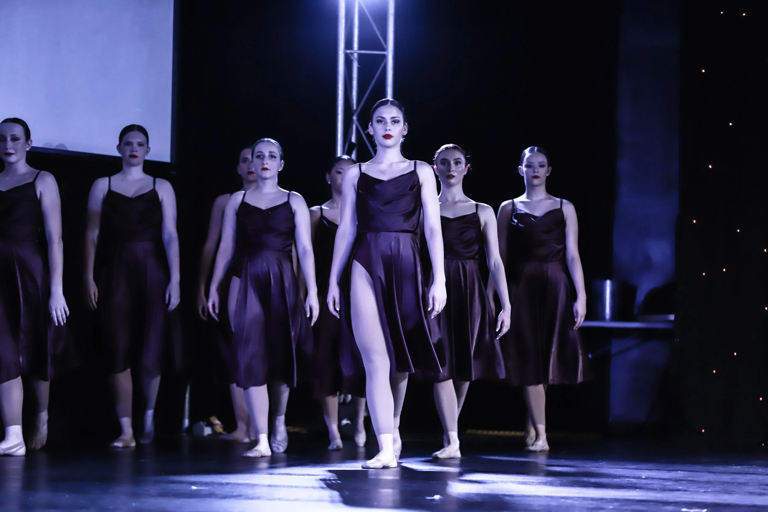 A group of ballet dancers in black dresses performing on stage, with one dancer in front, under stage lighting.