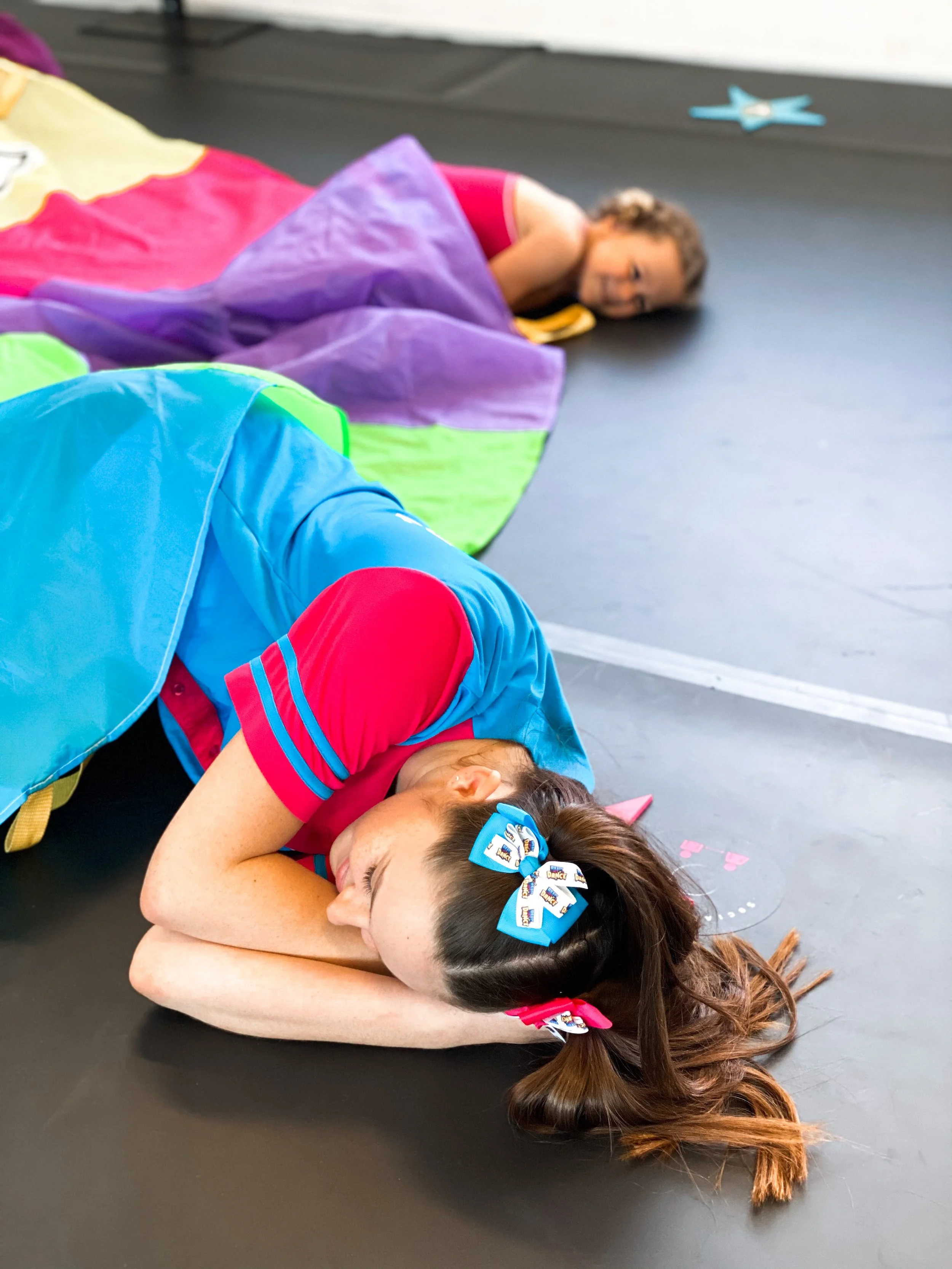 Two young girls lying on gym floor, resting with eyes closed, in colorful leotards and hair tied with bows.