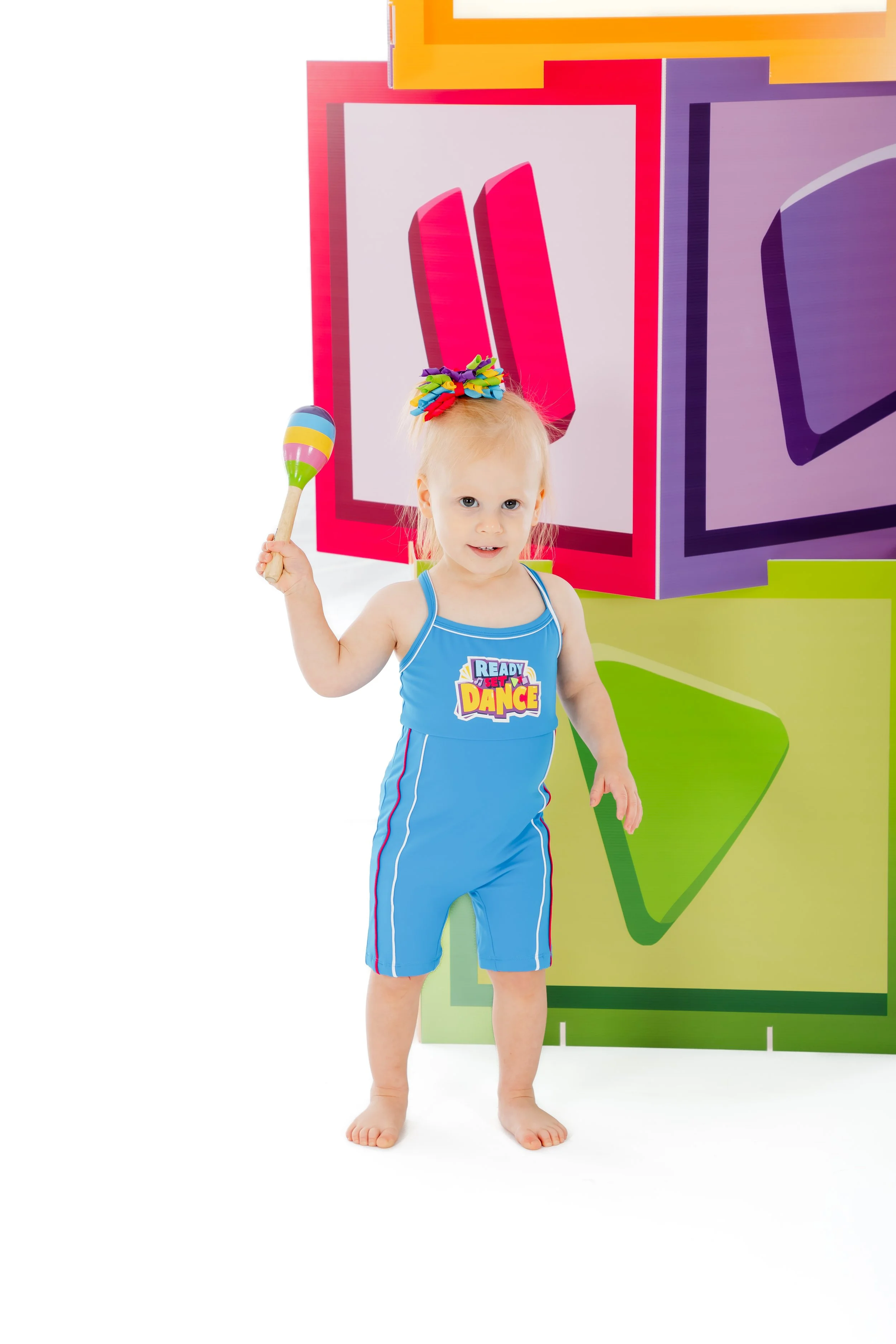 A young girl with blonde hair in a blue athletic outfit holding a colorful maraca stands in front of large, colorful geometric cube shapes.
