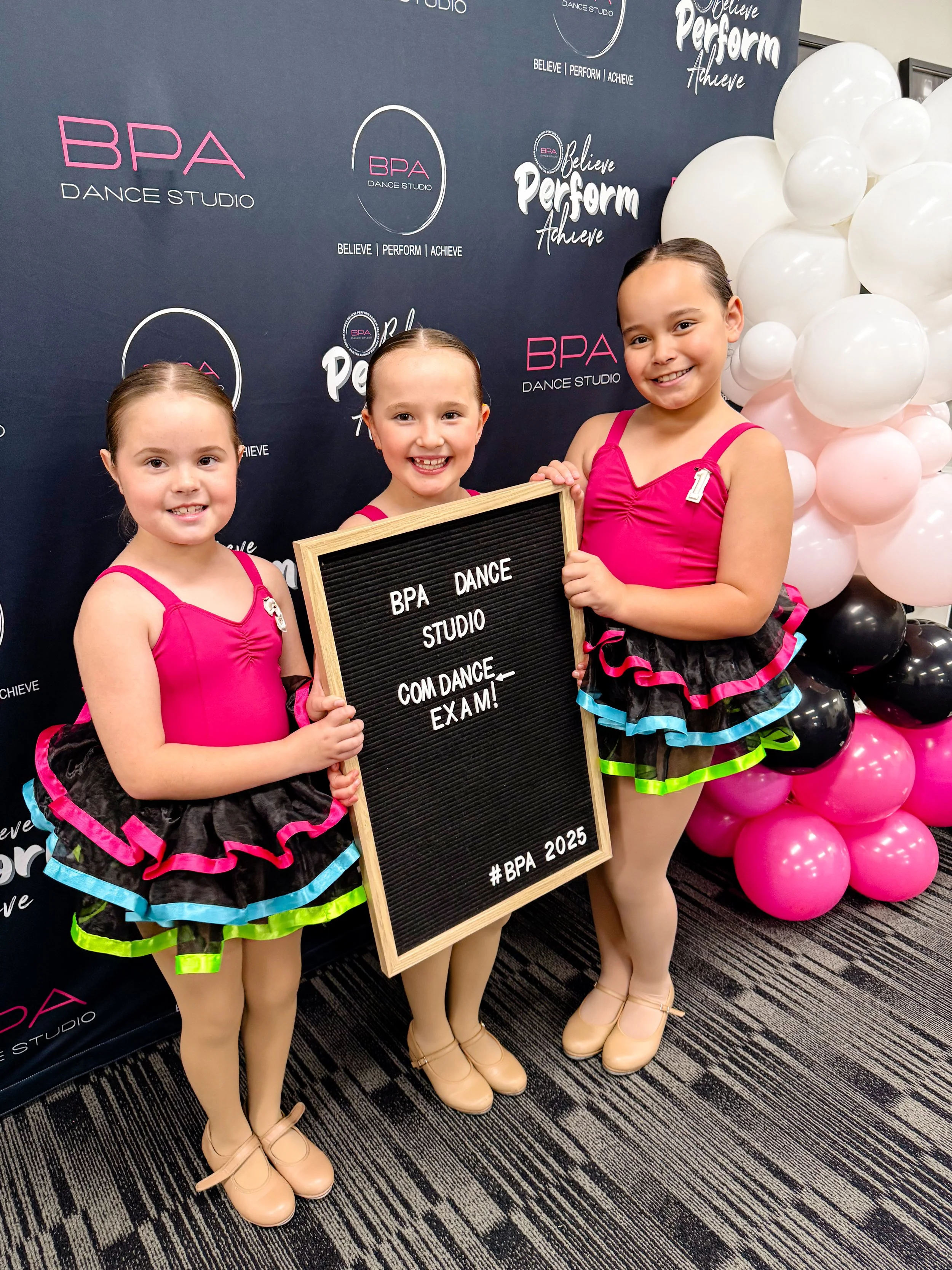 Three young girls in pink and black dance costumes holding a sign at a dance studio event, with a backdrop featuring the studio's logo and balloons.